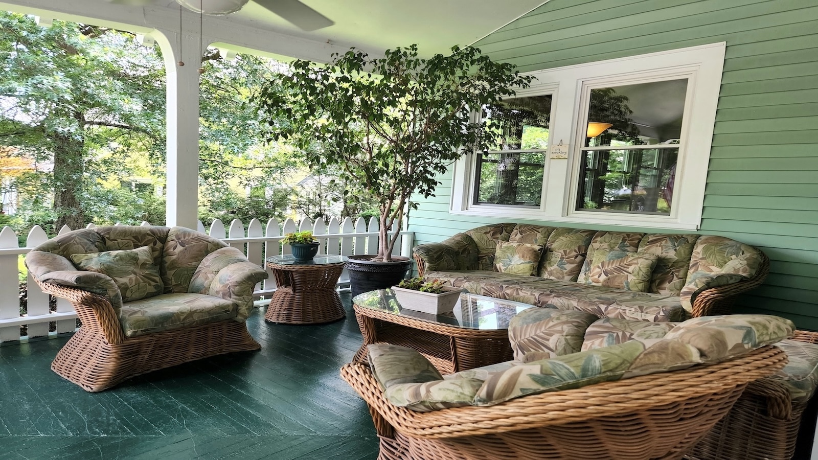 Covered painted porch with white railing, cushioned wicker furniture and stands, planted tree in pot in corner