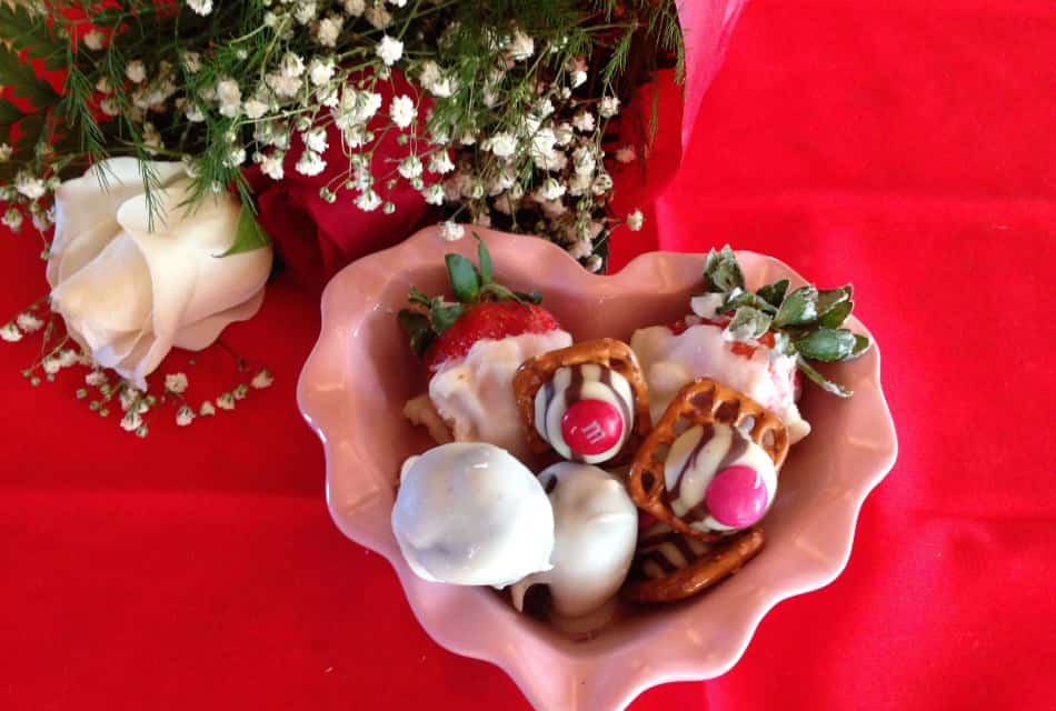 Small pink heart shaped bowl full of white chocolate covered strawberries and candies next to a white and red rose