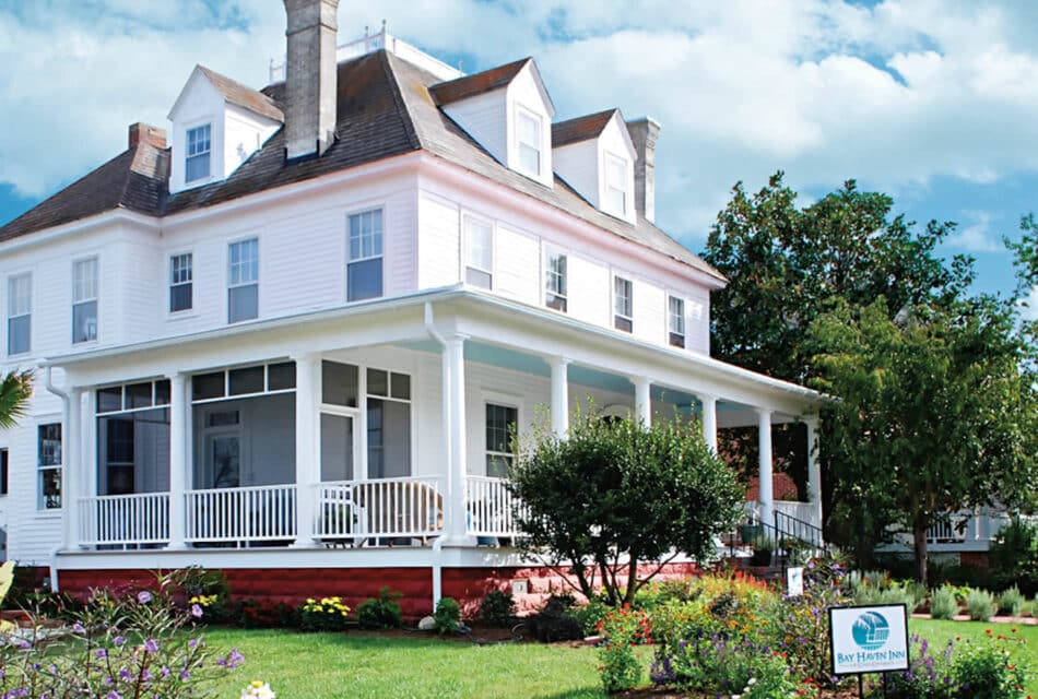 Exterior view of property painted all white with wrap around porch surrounded by green grass, shrubs, flowers and trees