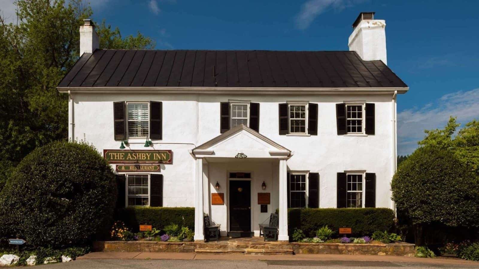 Exterior view of property painted white with black shutters and surrounded by green shrubs, bushes, and trees