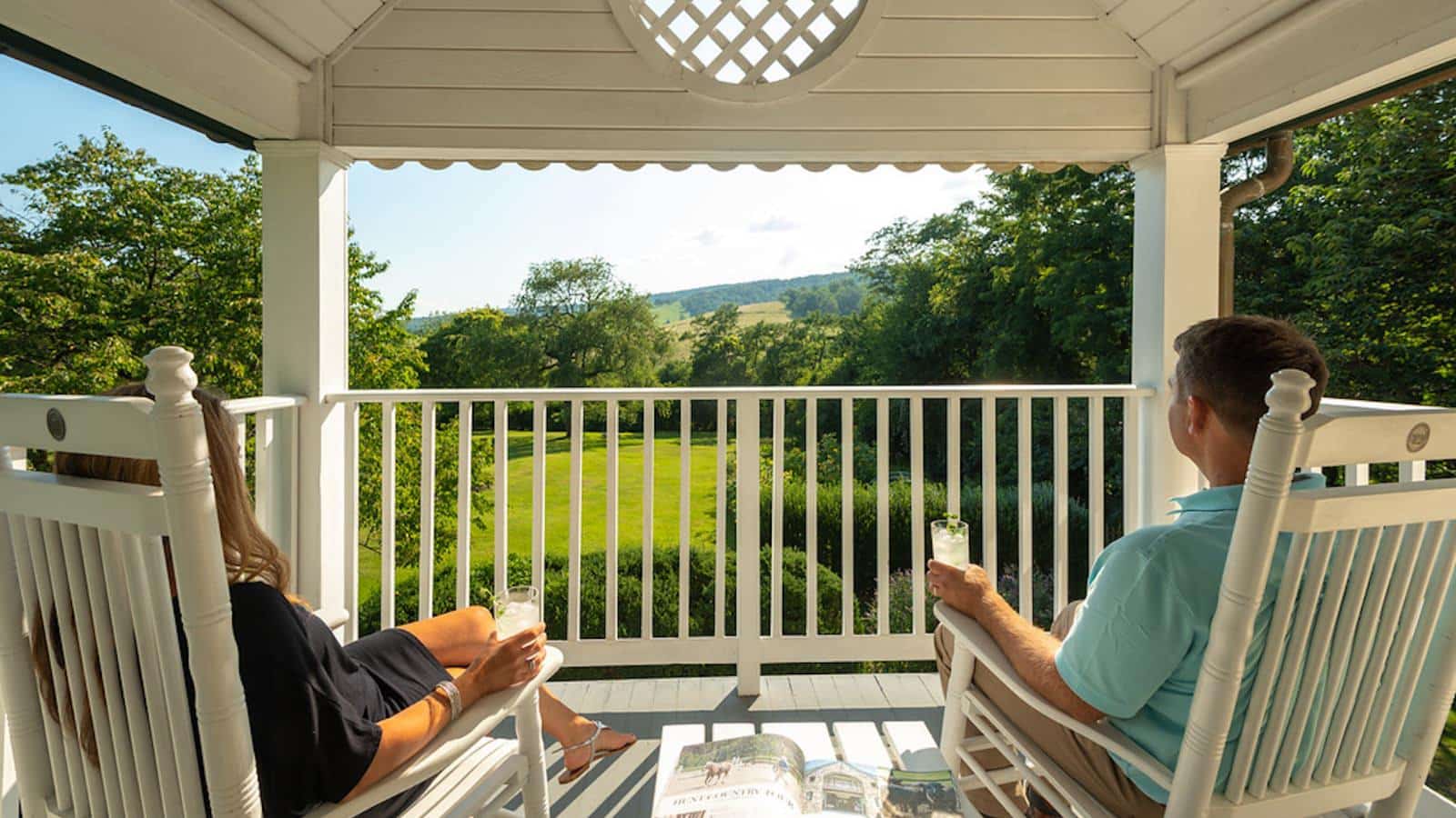 Covered patio with white wooden rocking chairs filled with people looking out over the green grass, green trees, and rolling hills