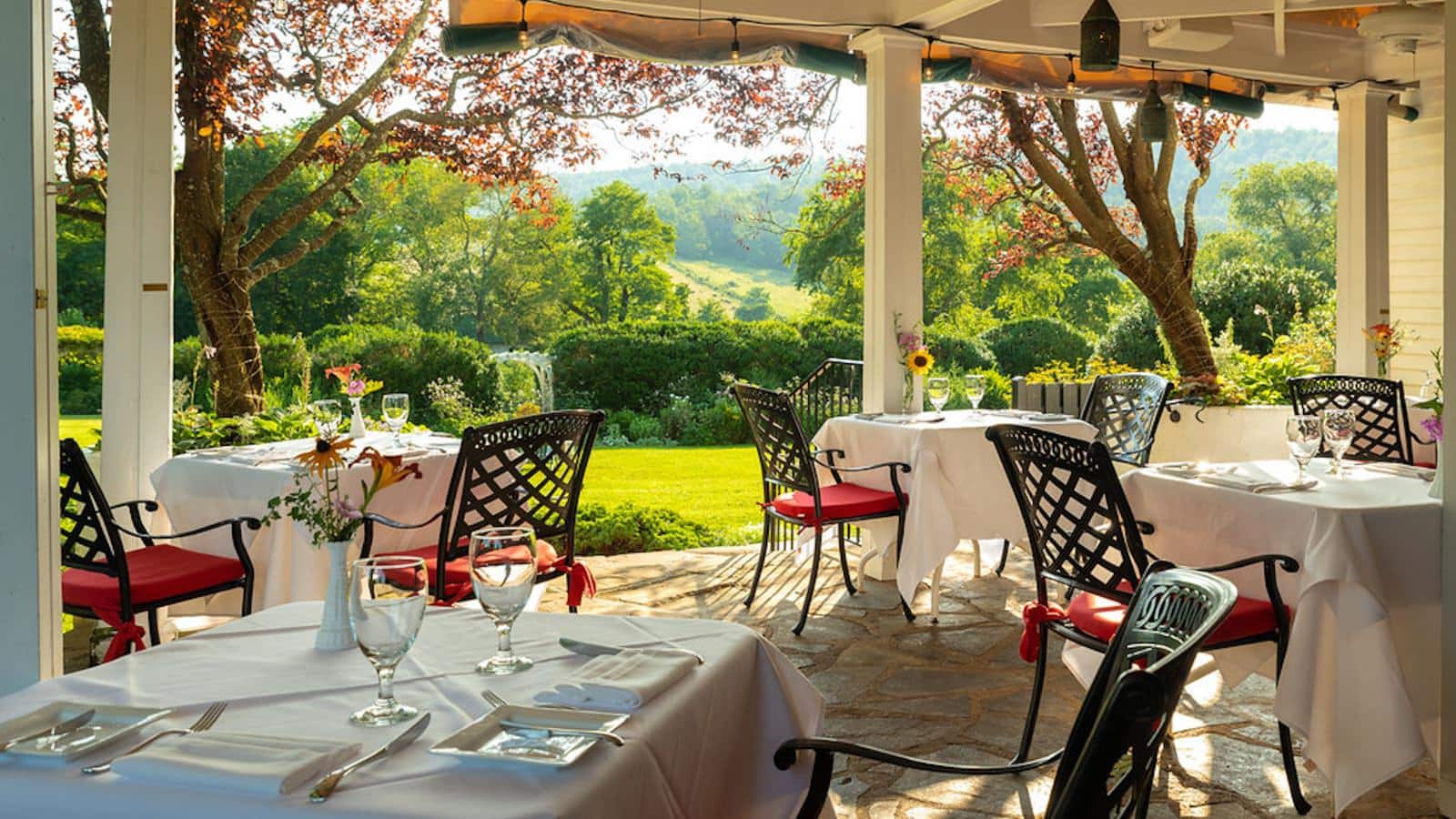 Covered patio with multiple tables with white tablecloths and place settings, wrought iron chairs with red cushions and view of green grass, bushes, trees, and rolling hills in the background