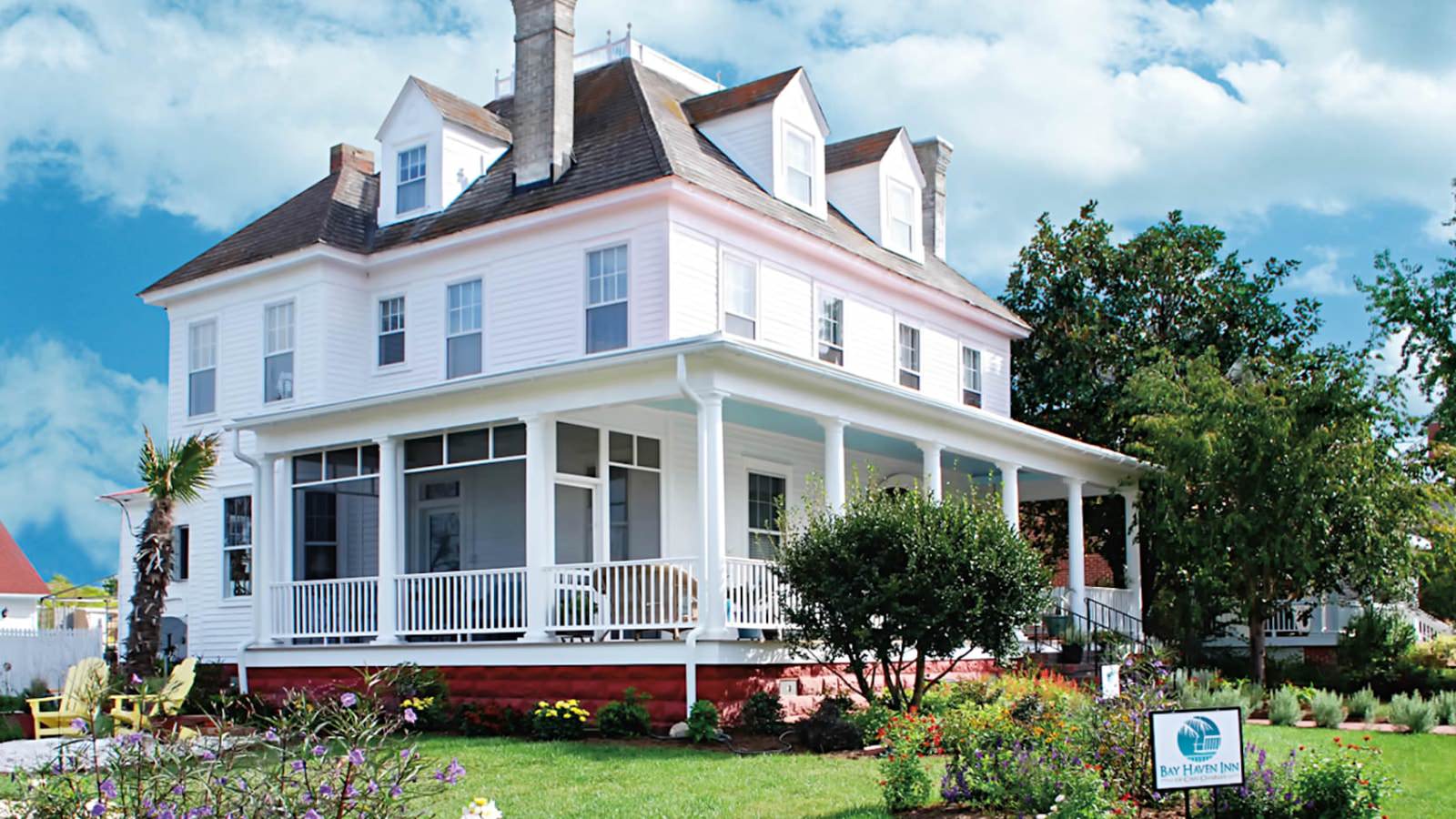 Exterior view of property painted all white with wrap around porch surrounded by green grass, shrubs, flowers and trees