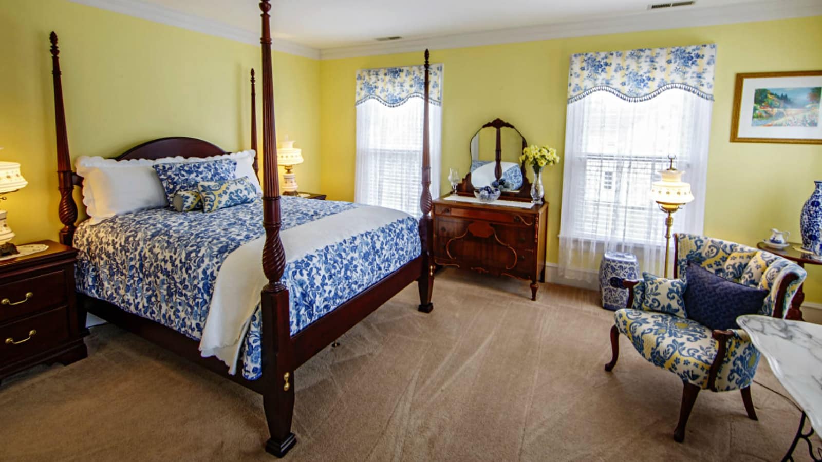 Bedroom with yellow walls, carpeting, dark wood four poster bed, blue multicolored bedding, dark wooden dresser, and upholstered and wooden antique chair