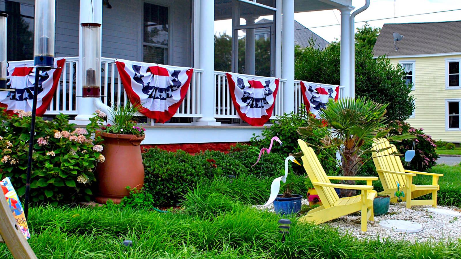 Exterior view of property's front porch with white columns and Independence Day flags on the railing, green bushes, green grass, and yellow Adirondack chairs on white gravel patio area