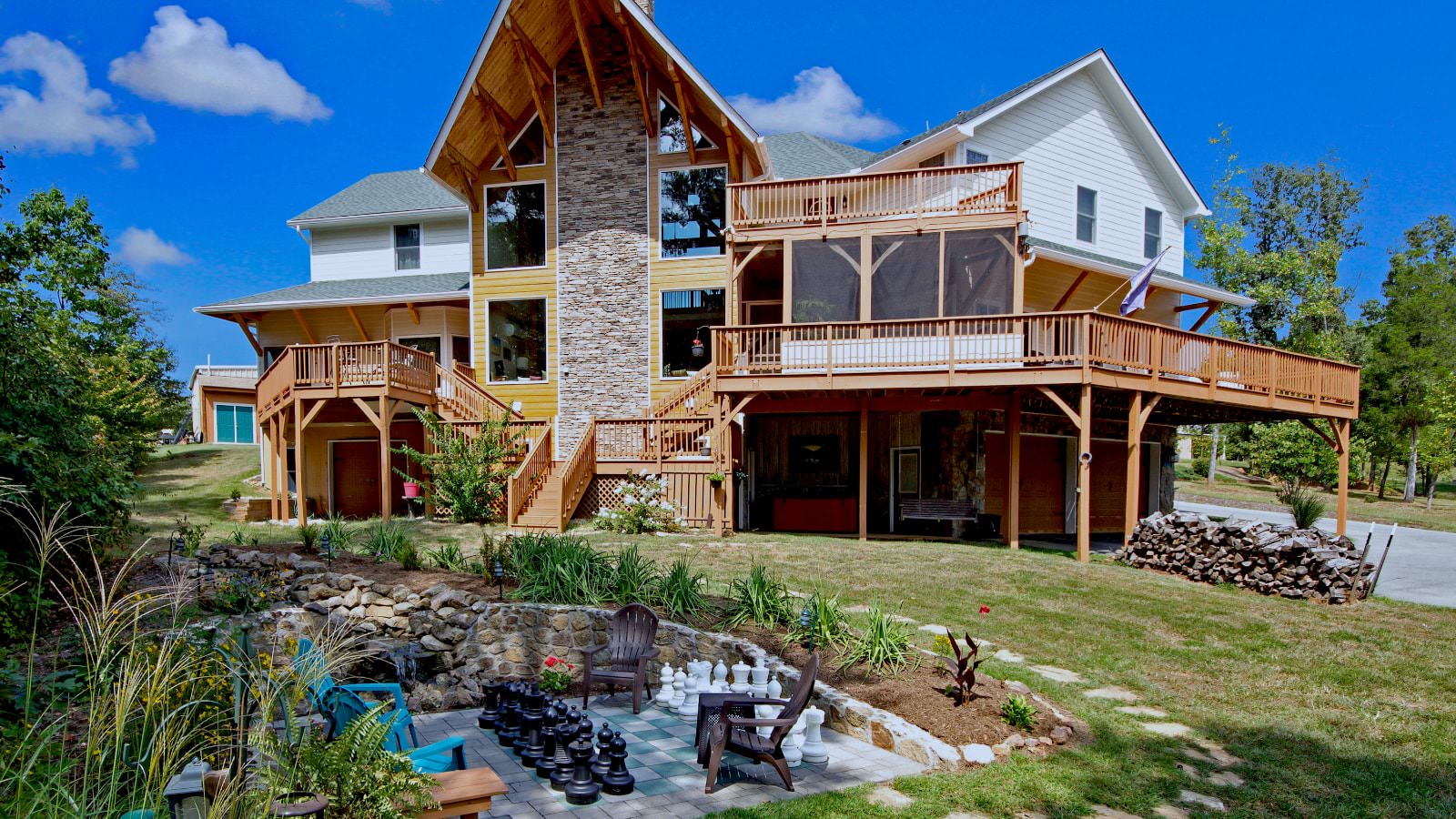 Exterior view of a three level property with large stone fireplace, wrap around porch, backyard patio chess, Adirondack chairs, and flower garden