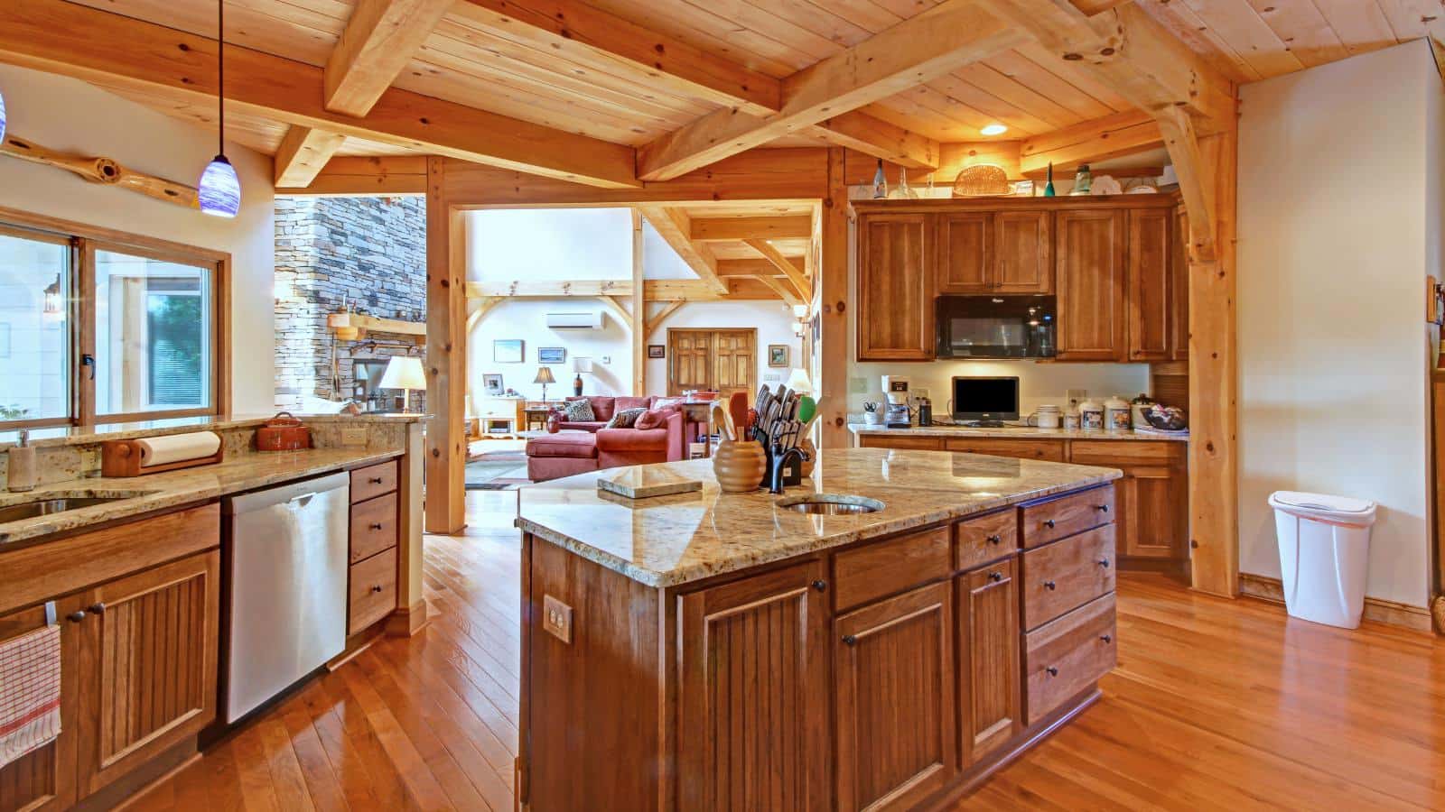 Large kitchen with wooden cabinets, marble countertops, white walls, wooden ceiling, and view into great room