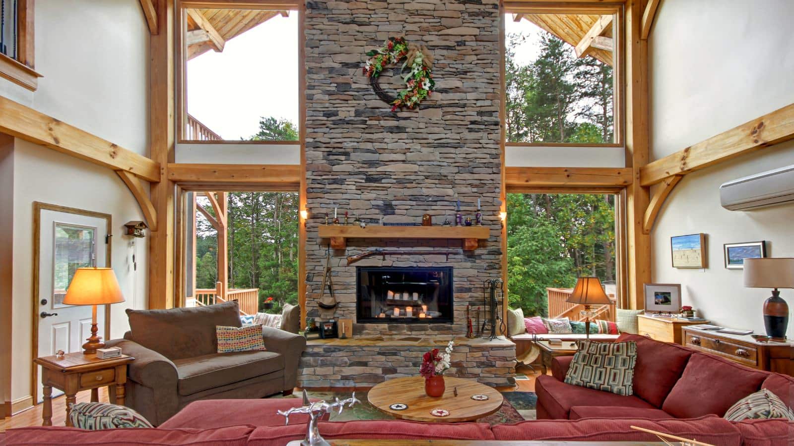 Great room with large stone fireplace, large red upholstered sectional, oversized brown upholstered chair, and large windows with views of green trees