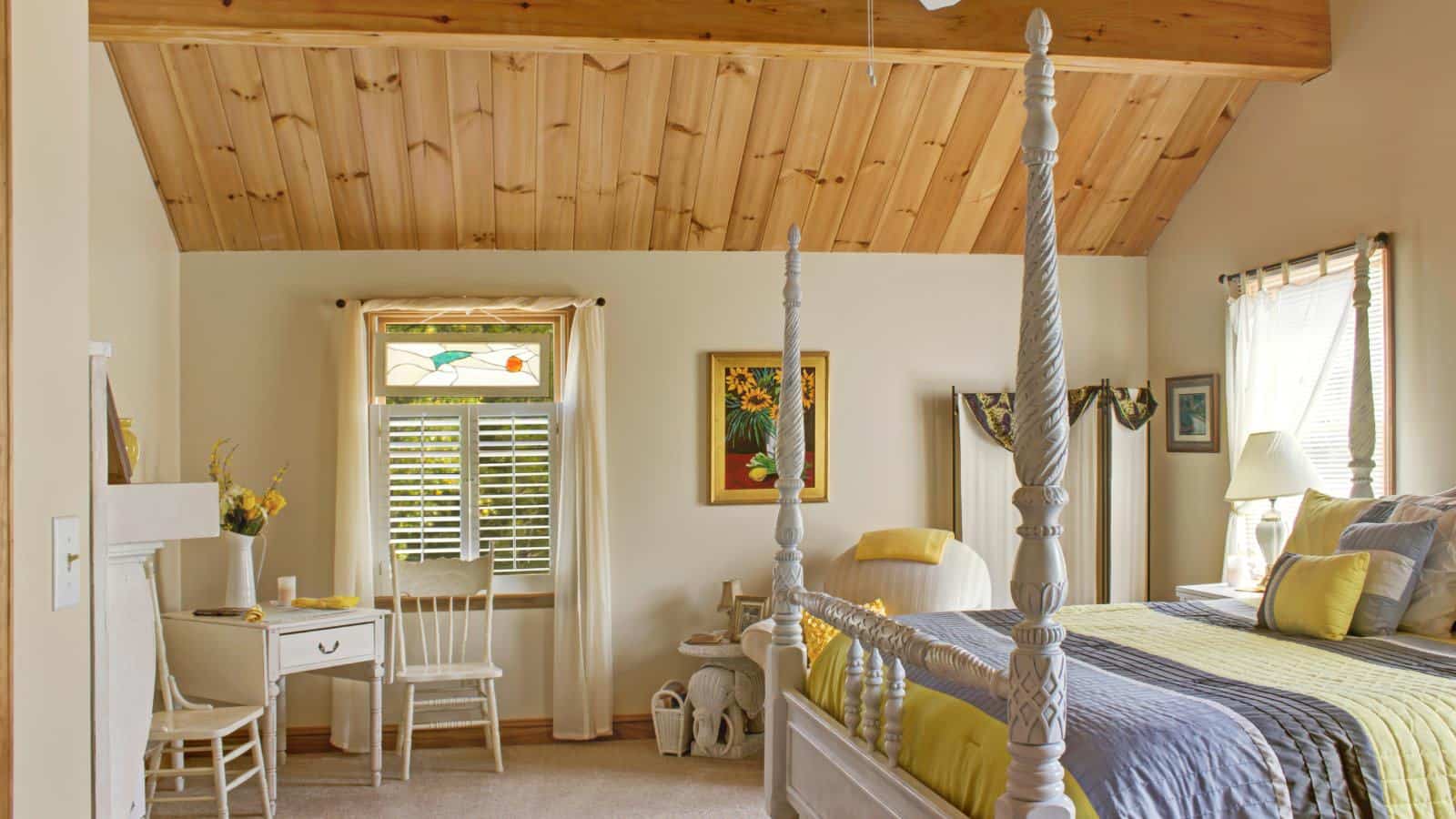 Bedroom with cream walls, wooden ceiling, white four poster bed, and sitting area with white wooden chairs