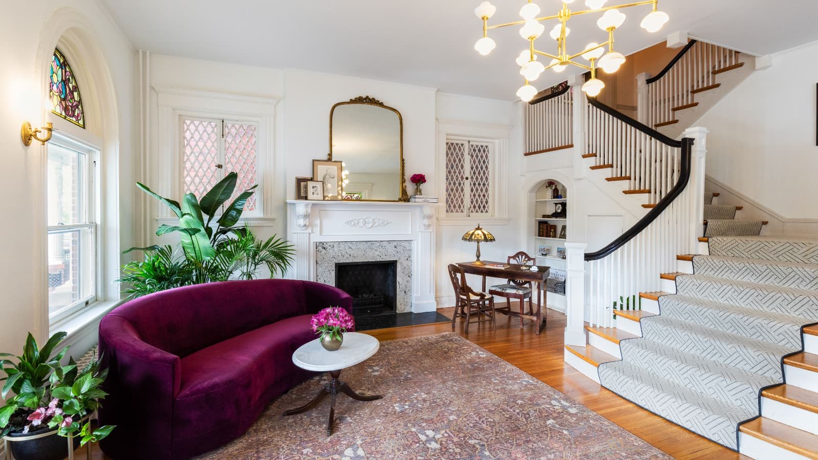 Living room with white walls, hardwood flooring, dark purple upholstered sofa, marble coffee table, desk with two chairs, fireplace, and staircase up to the second floor