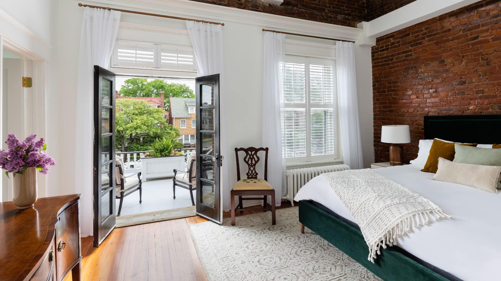Bedroom with white walls and red brick accent wall behind bed with white bedding, hardwood flooring, and open double doors to balcony