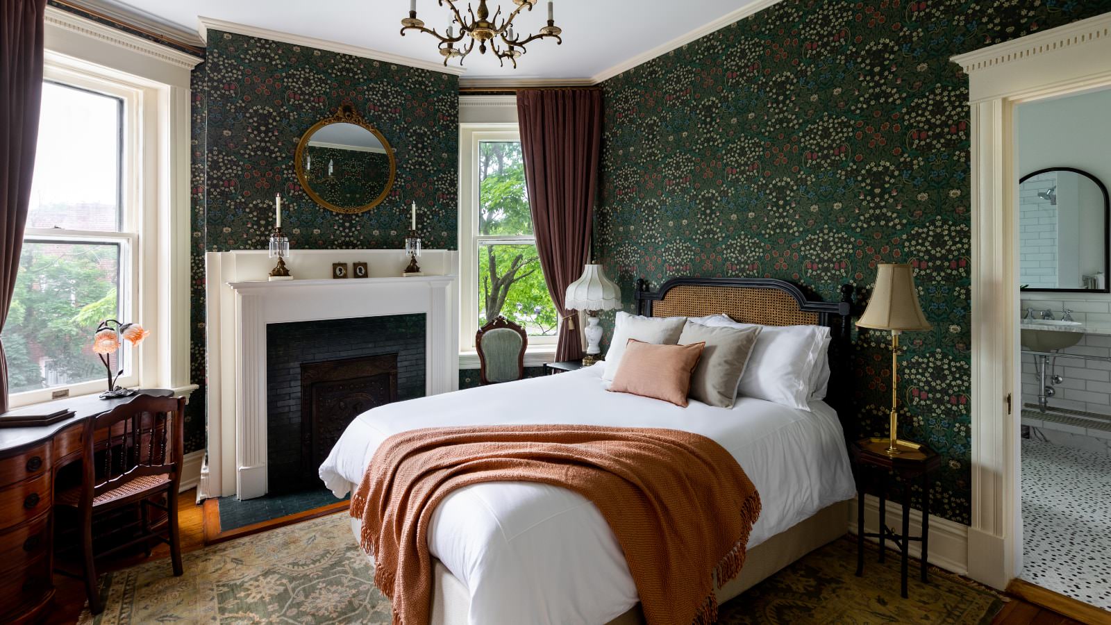 Bedroom with dark green ornate wallpaper, white trim, hardwood flooring, bed with white bedding, wooden antique desk, fireplace, and large windows