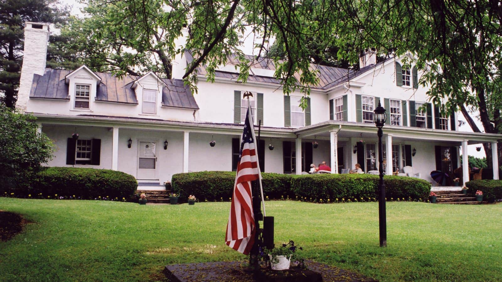 Exterior view of property painted white with dark green shutters surrounded by green grass, shrubs, and trees