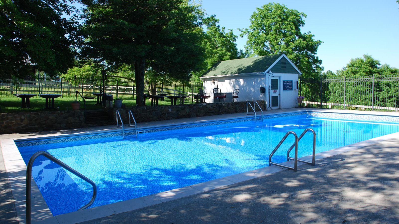 Pool surrounded by wrought iron fencing, pool house, tables, and green trees