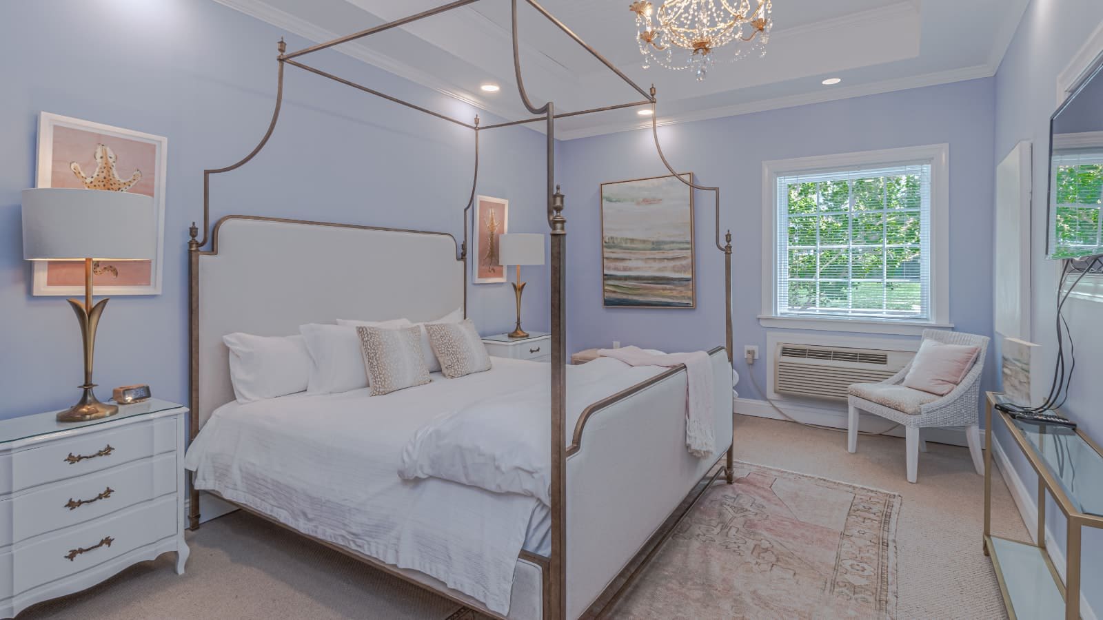 Bedroom with light lavender walls, carpeting, metal four poster bed, white bedding, white dressers, and white wicker chair