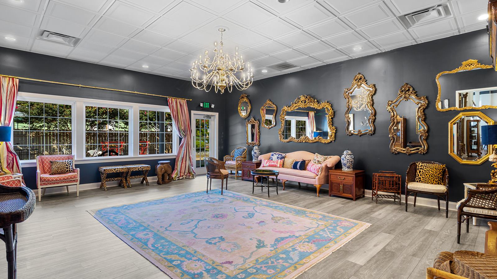 Large room with dark walls, hardwood flooring, large chandelier, large area rug, multiple ornate mirrors, and multicolored sofas and tables set up around the edge of the room