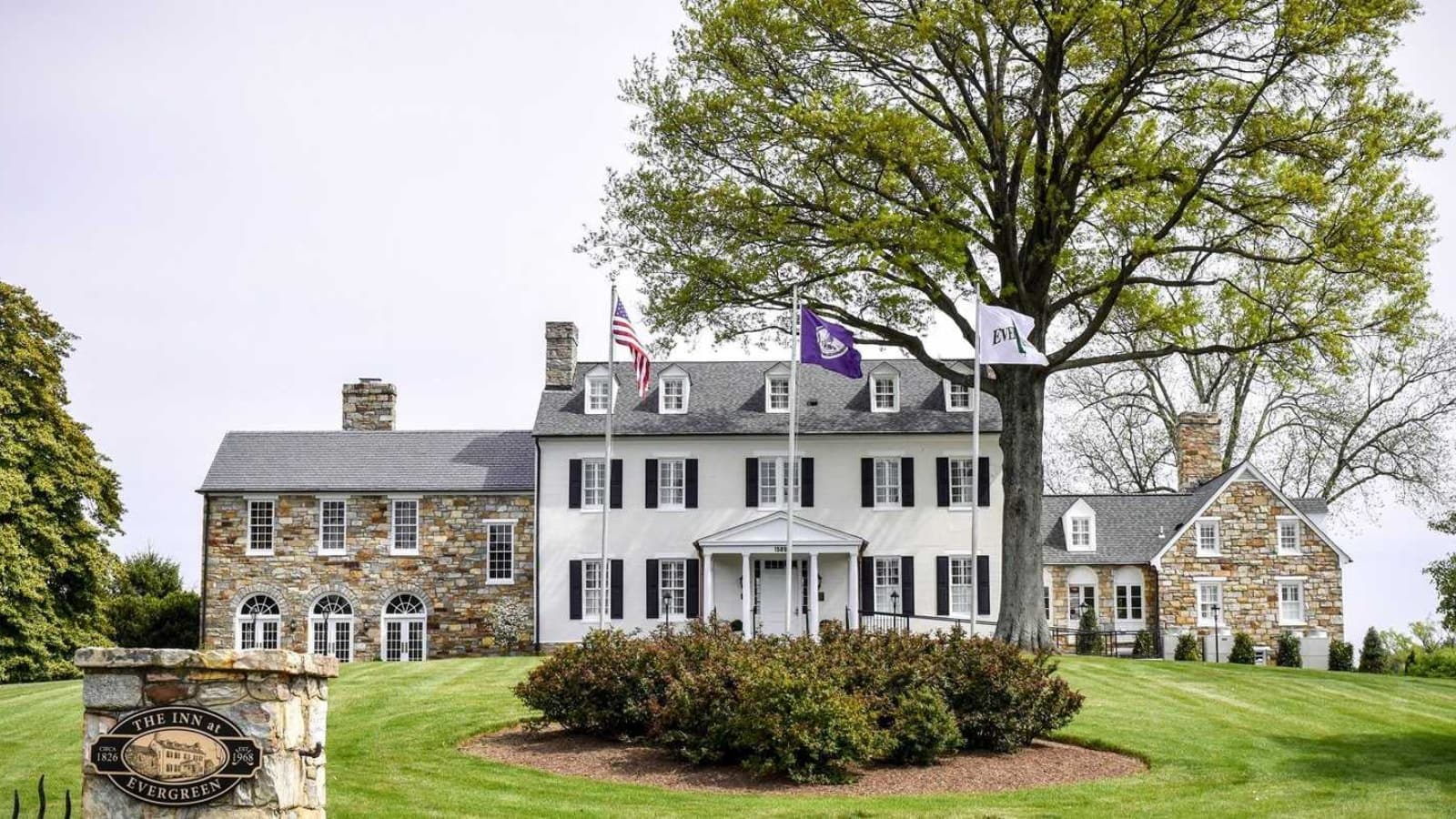 Exterior view of the property painted white with black shutters and stone brick surrounded by green grass, bushes, and trees