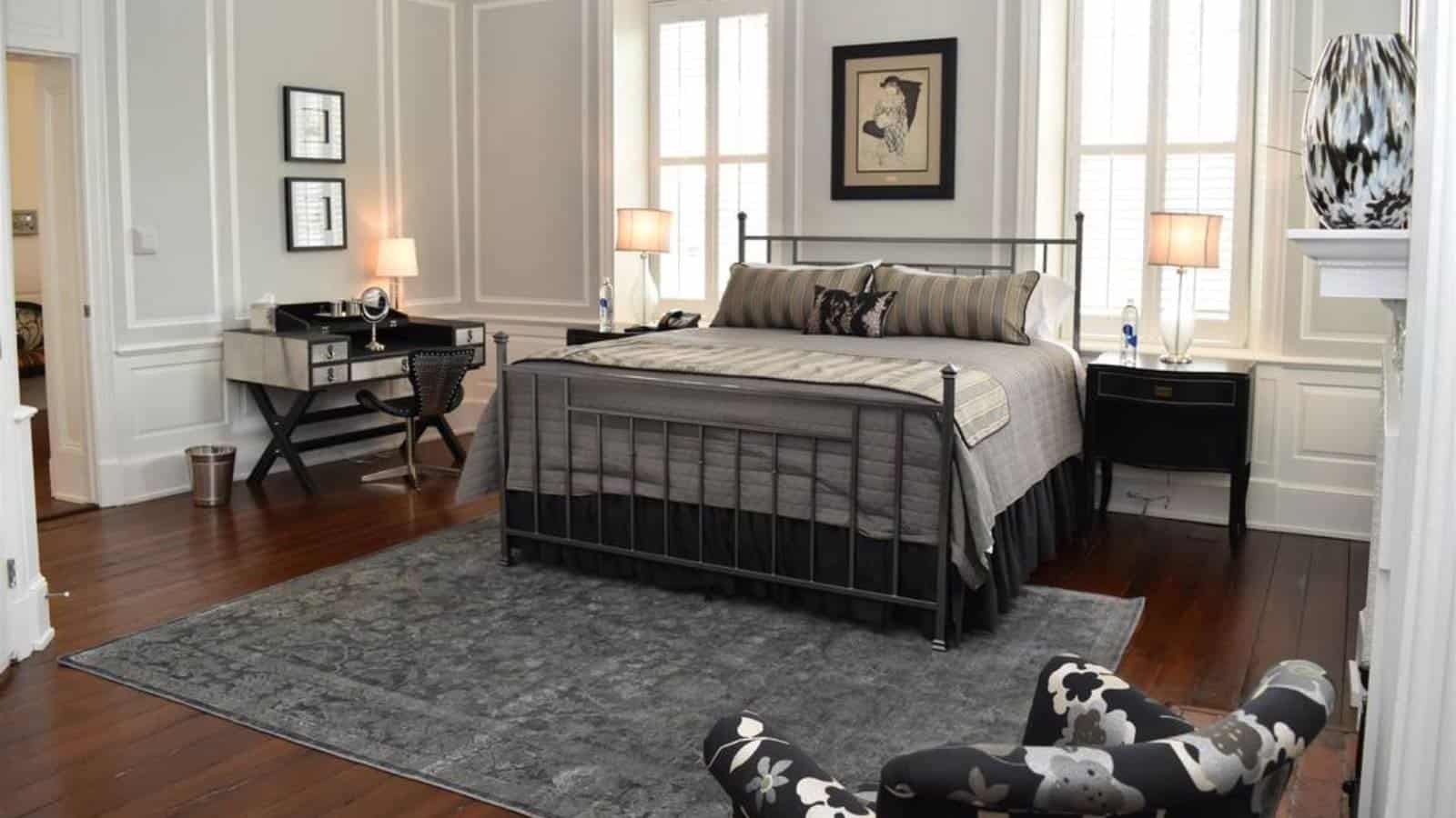 Bedroom with paneled walls, hardwood flooring, gray wrought iron bed with gray bedding, desk, and upholstered arm chair
