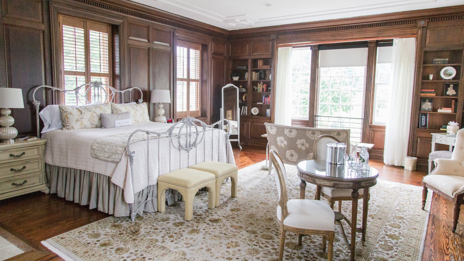 Bedroom with dark wooden wall paneling, hardwood flooring, white wrought iron bed with white bedding, large windows, and sitting area