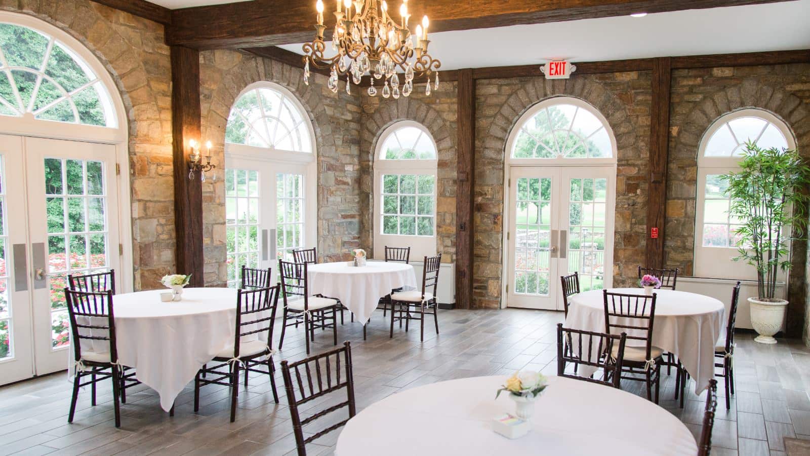 Large event area with stone brick walls, hardwood flooring, wooden tables with white tablecloths, wooden chairs, and large chandelier