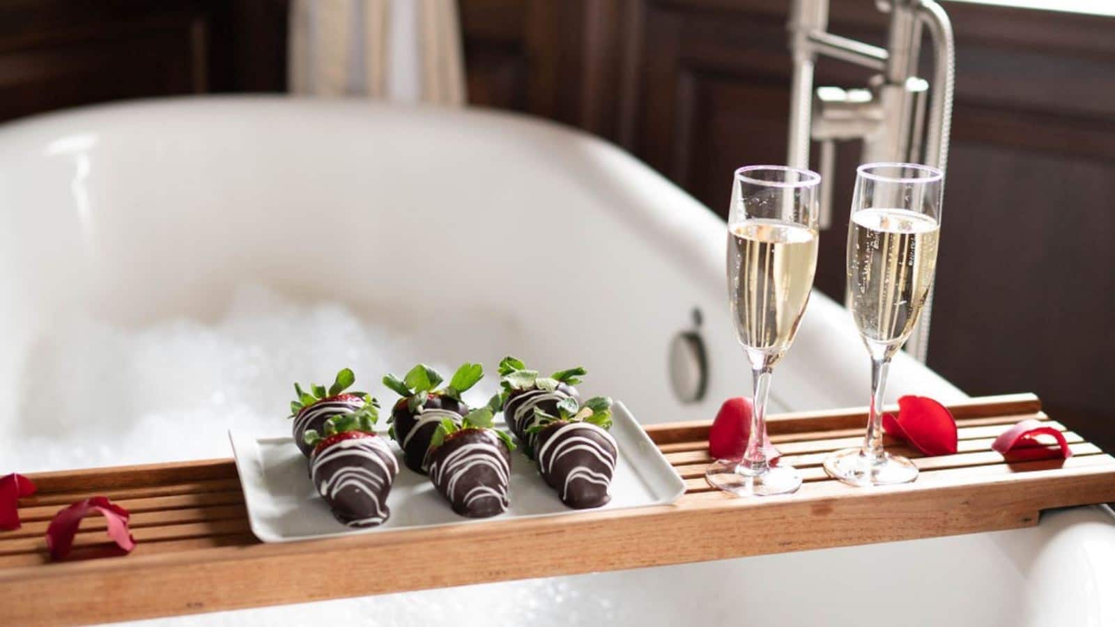 Close up view of small wooden tray across white claw foot tub with chocolate covered strawberries and glasses of Champagne