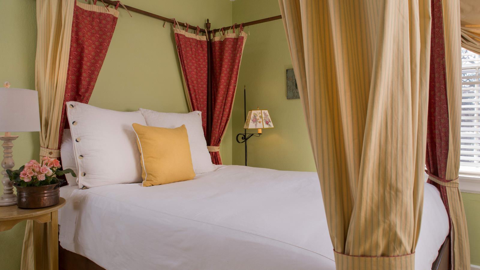 Bedroom with light lime green walls, four poster bed with yellow and red curtains and white bedding, antique floor lamp, and wooden side table