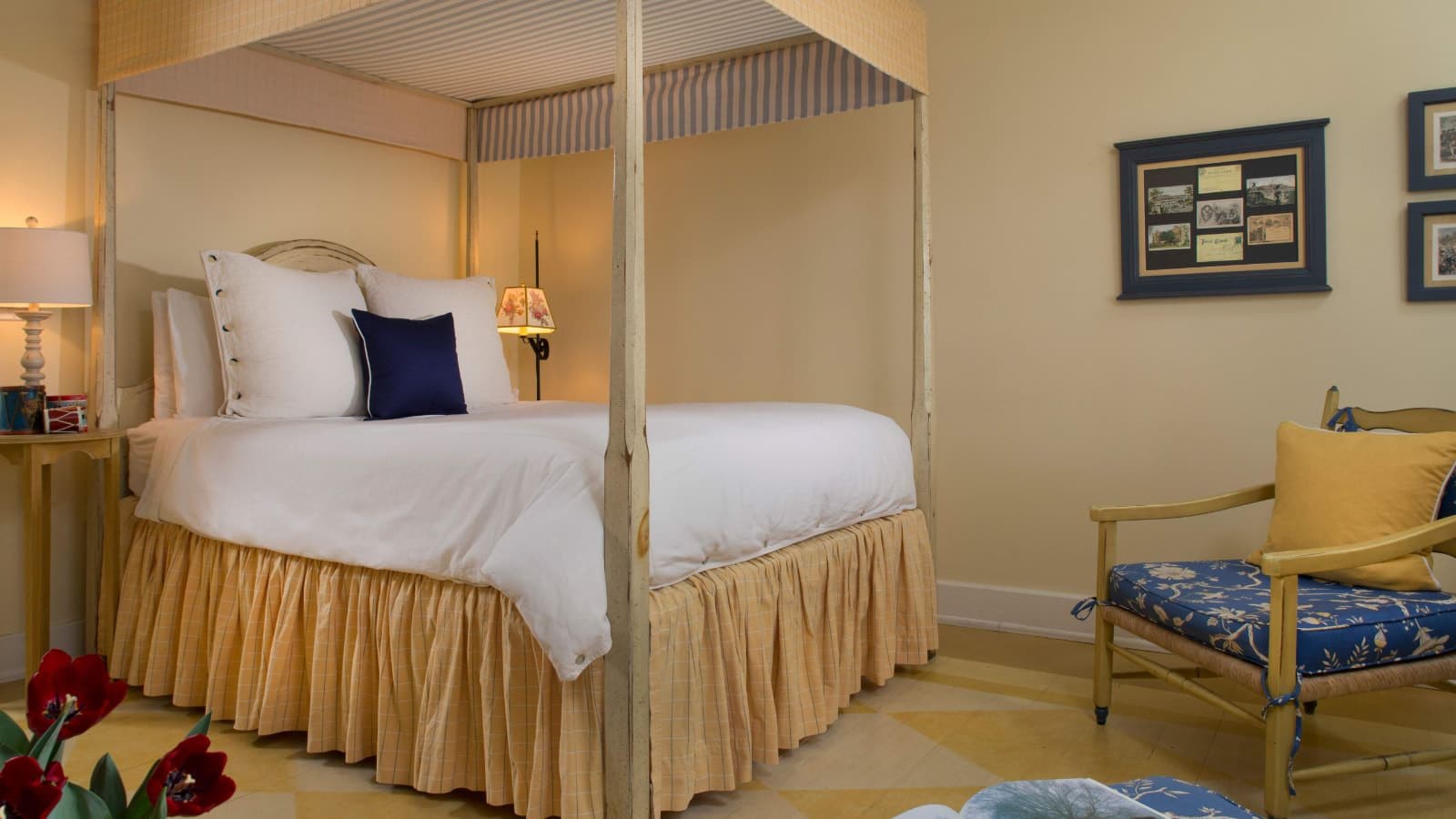 Bedroom with cream walls and hardwood flooring, four poster bed with canopy and white bedding, antique floor lamp, wooden side table, and wooden chair with blue floral cushion