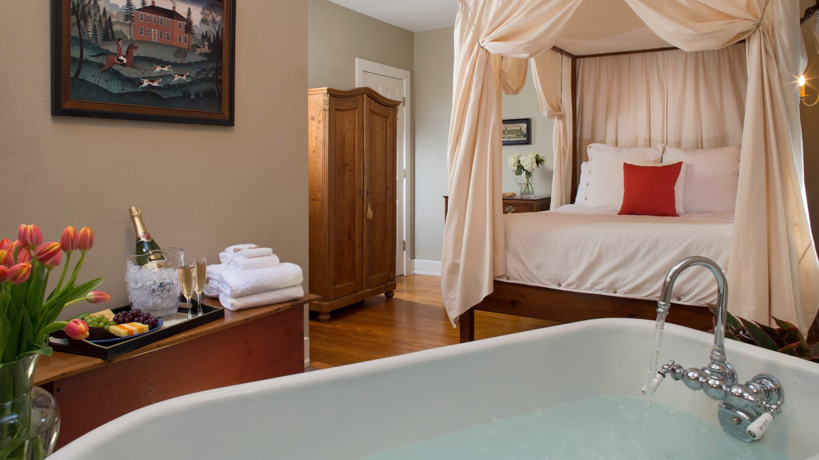 Bedroom with light tan walls, hardwood flooring, four poster bed with canopy and white bedding, and claw-foot tub