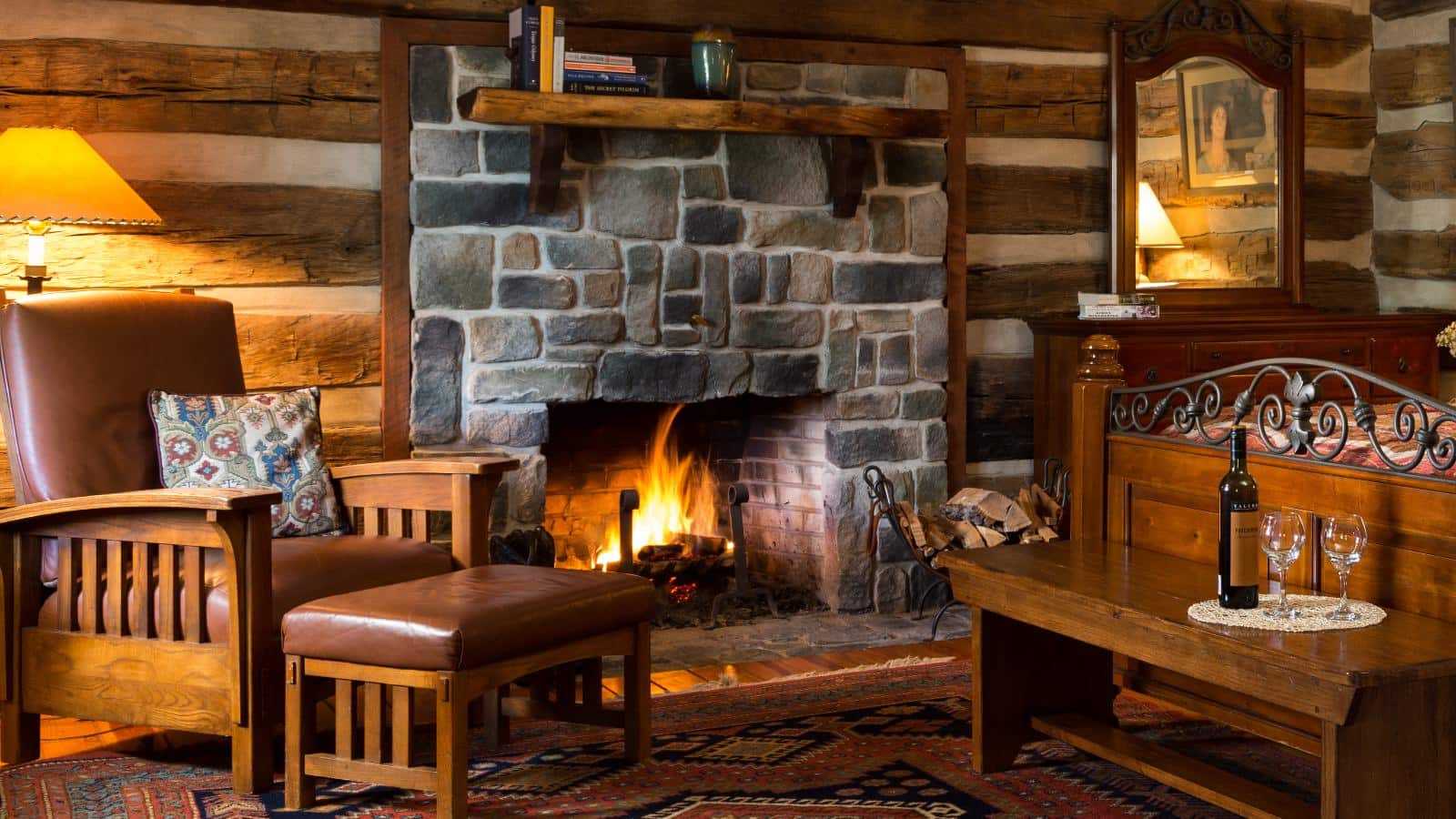 Bedroom with log cabin walls, stone fireplace, hardwood flooring, dark wooden bed and dresser, and wooden chair with brown leather cushions