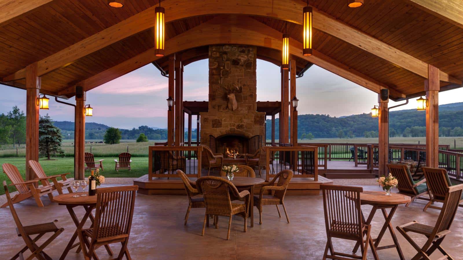 Large wooden covered patio with stone fireplace, wooden tables and chairs, and wooden Adirondack chairs with green grass and rolling hills in the background