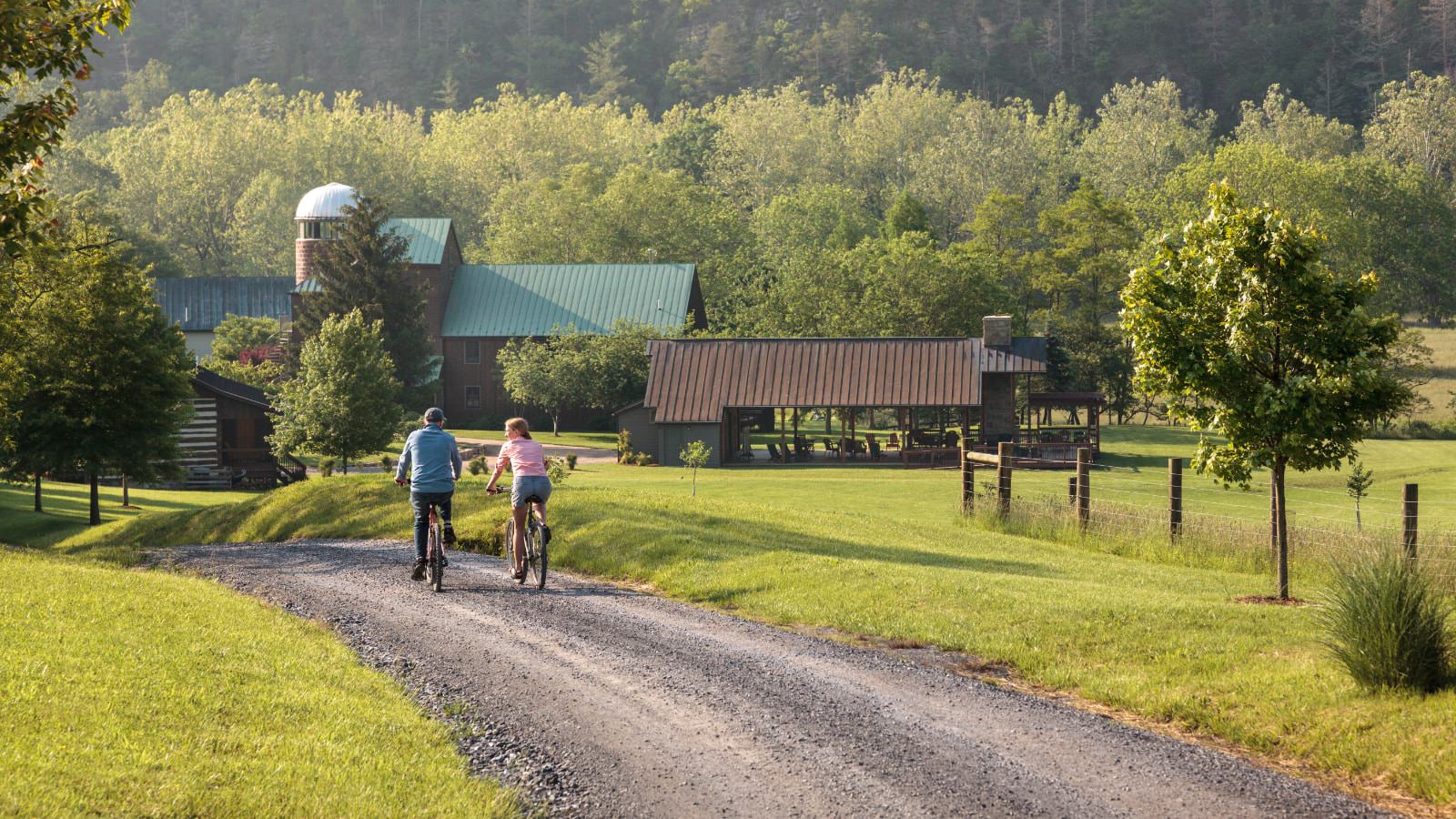 Two people riding bikes on gravel road with property and green trees in the background