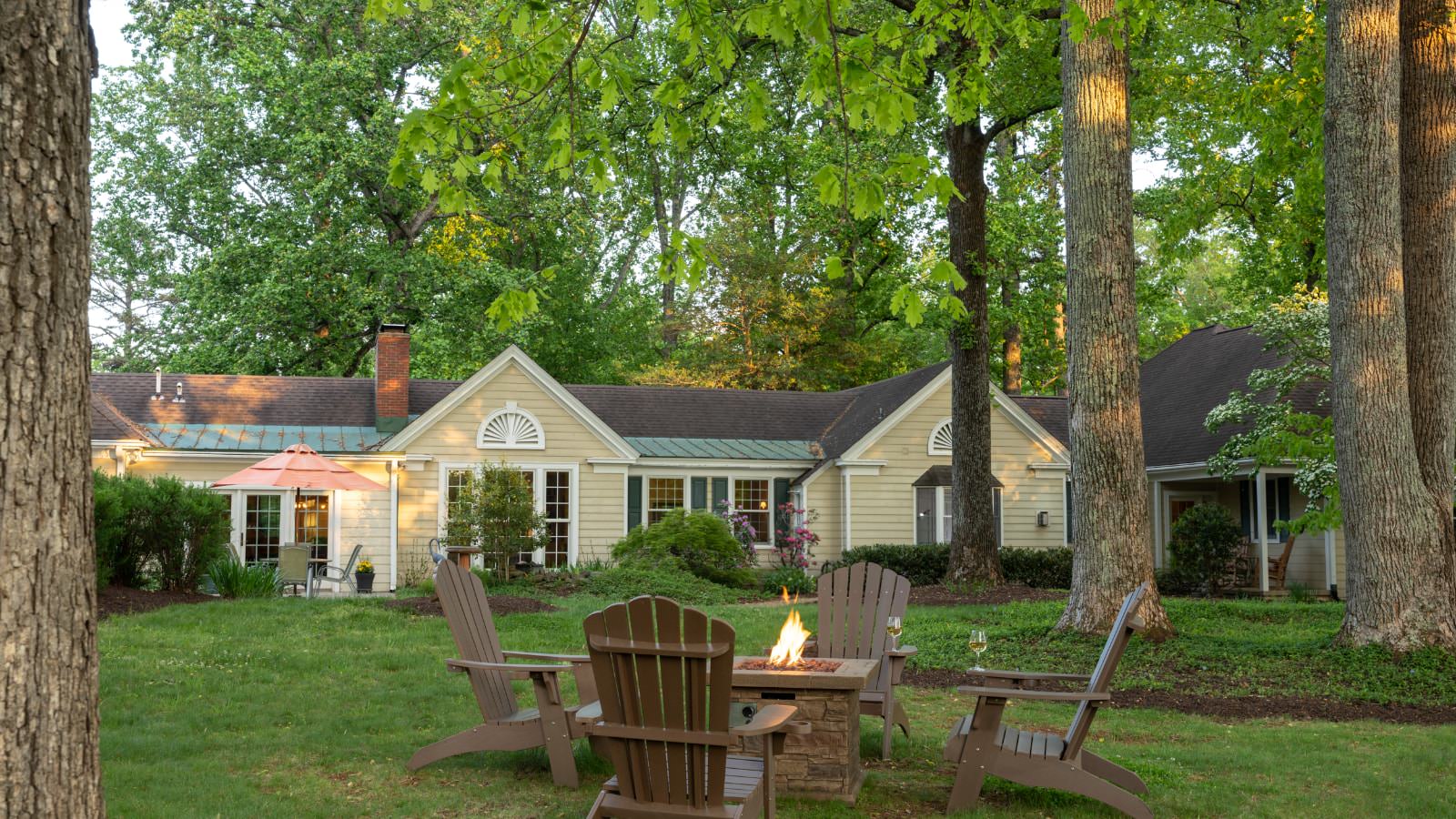 Exterior view of property painted yellow with white trim and firepit with four brown Adirondack chairs surrounded by green grass and very tall green trees