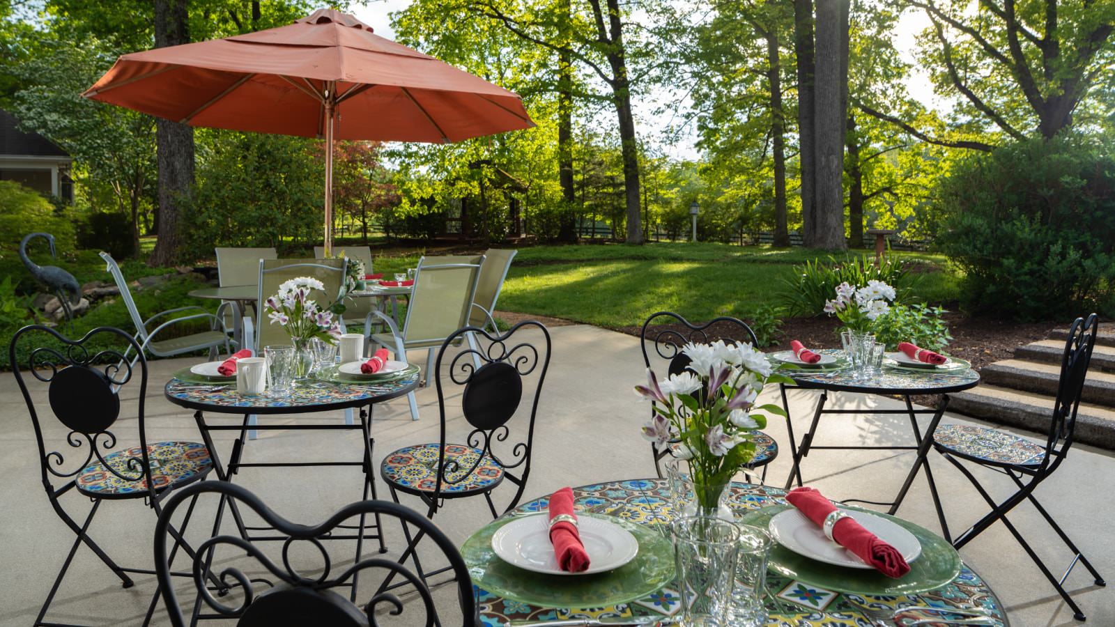 Concrete patio with black wrought iron tables and chairs and a patio dining table with chairs and brown umbrella with tall trees in the background