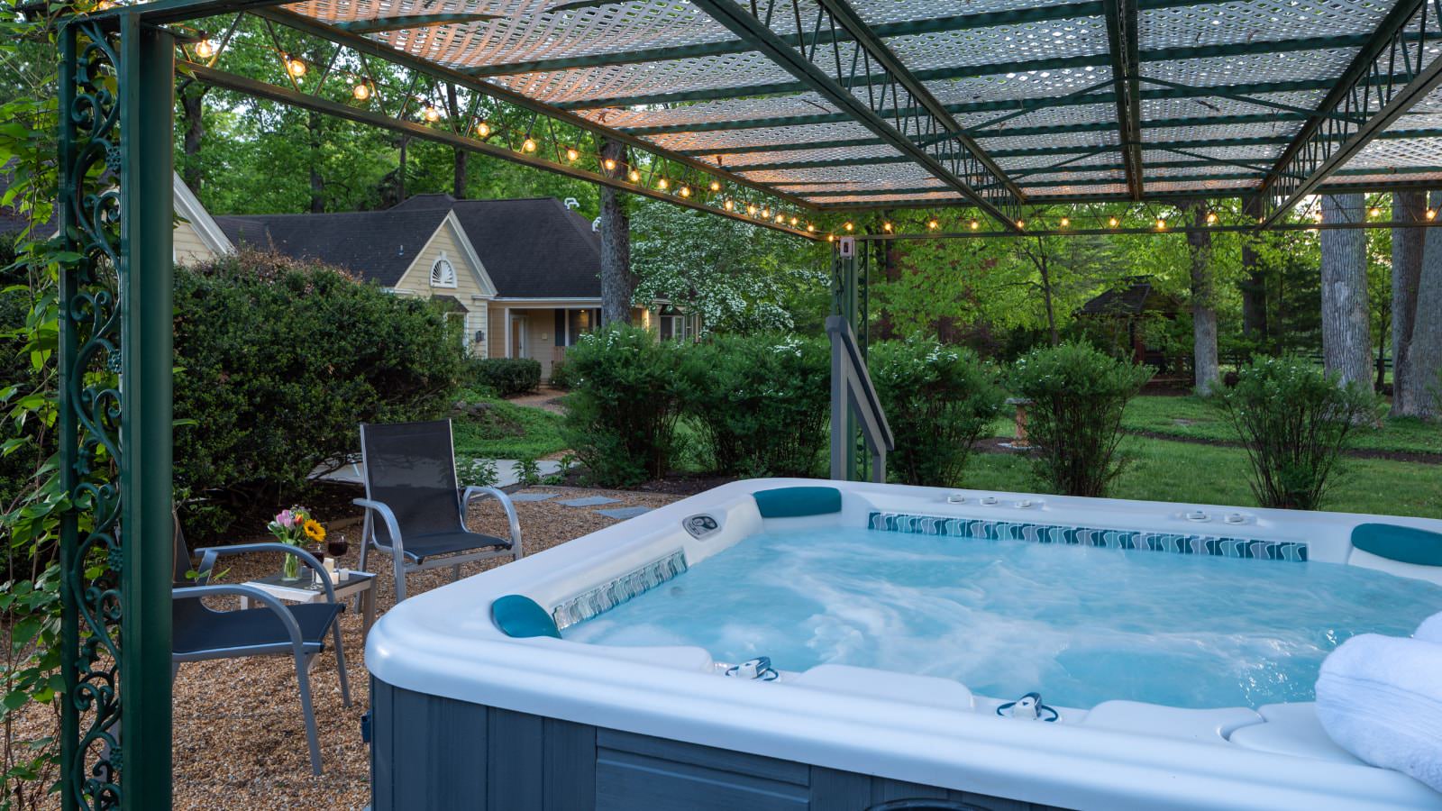 Outdoor hot tub under a green wrought iron covering decorated with lights and two patio chairs and a small table near by with green bushes in the background