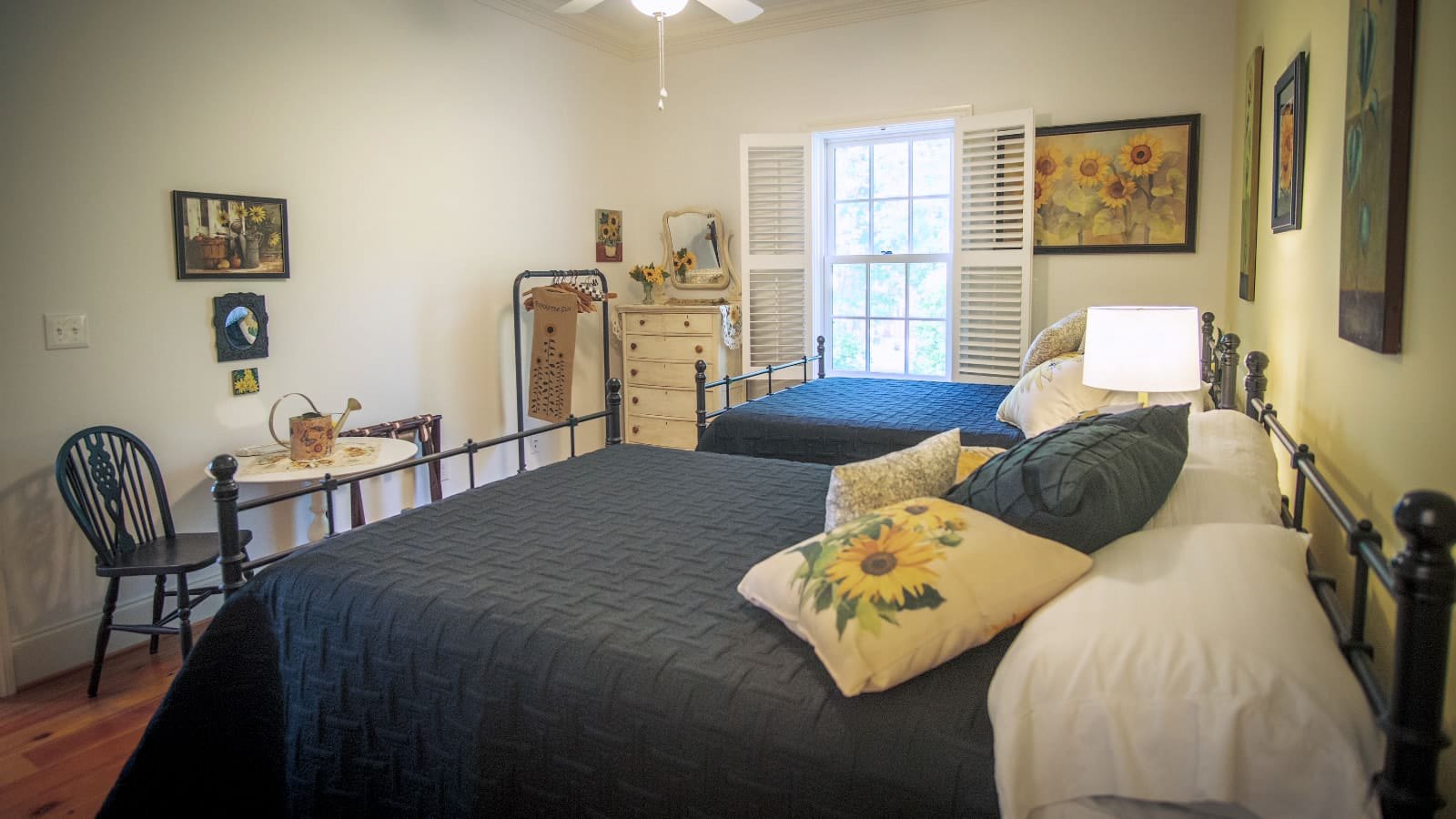 Bedroom with white walls, hardwood flooring, two wrought iron beds with navy bedding, white wooden dresser, and sitting area