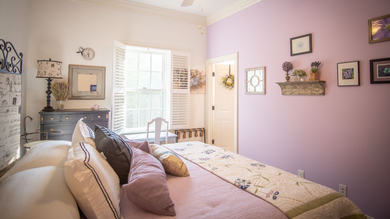 Bedroom with light purple and white walls, antique headboard, light purple bedding, dark wooden dresser, and white desk and chair
