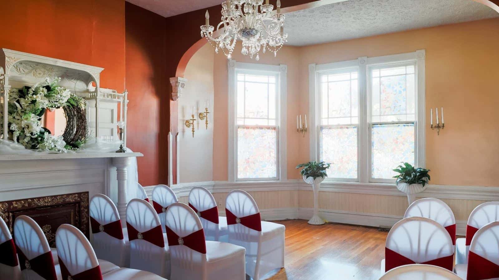 Large living room set up for a wedding with red and apricot walls, hardwood flooring, fireplace, chandelier, and white chairs with white and red fabric coverings