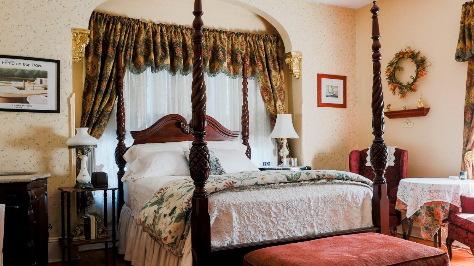 Bedroom with light colored wallpaper and walls, hardwood flooring, dark wooden four poster bed, white bedding, and sitting area