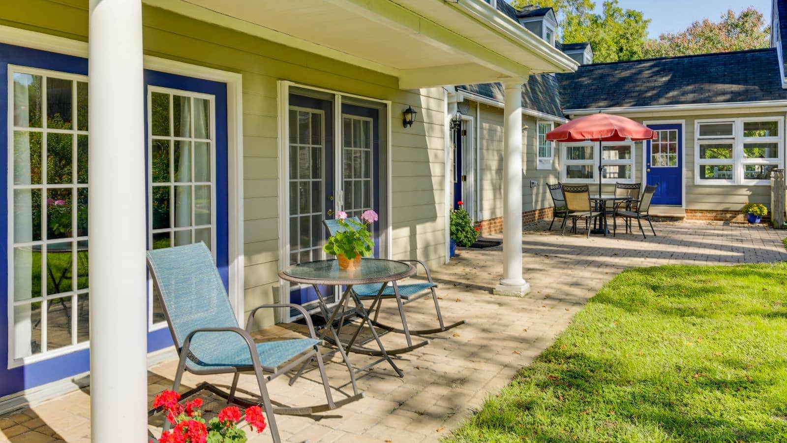 Exterior view of property painted light taupe with white trim and blue doors, paver patio with patio dining table and chairs and red umbrella and green grass