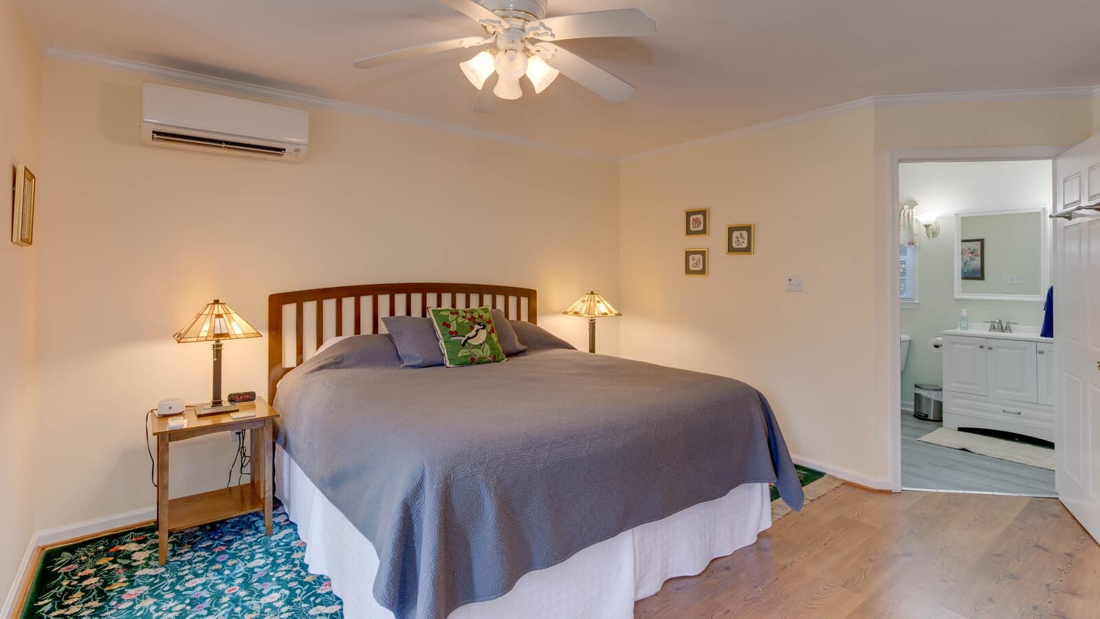 Bedroom with cream walls, hardwood flooring, wooden bed, gray bedding, wooden nightstands with lamps, and view into bathroom