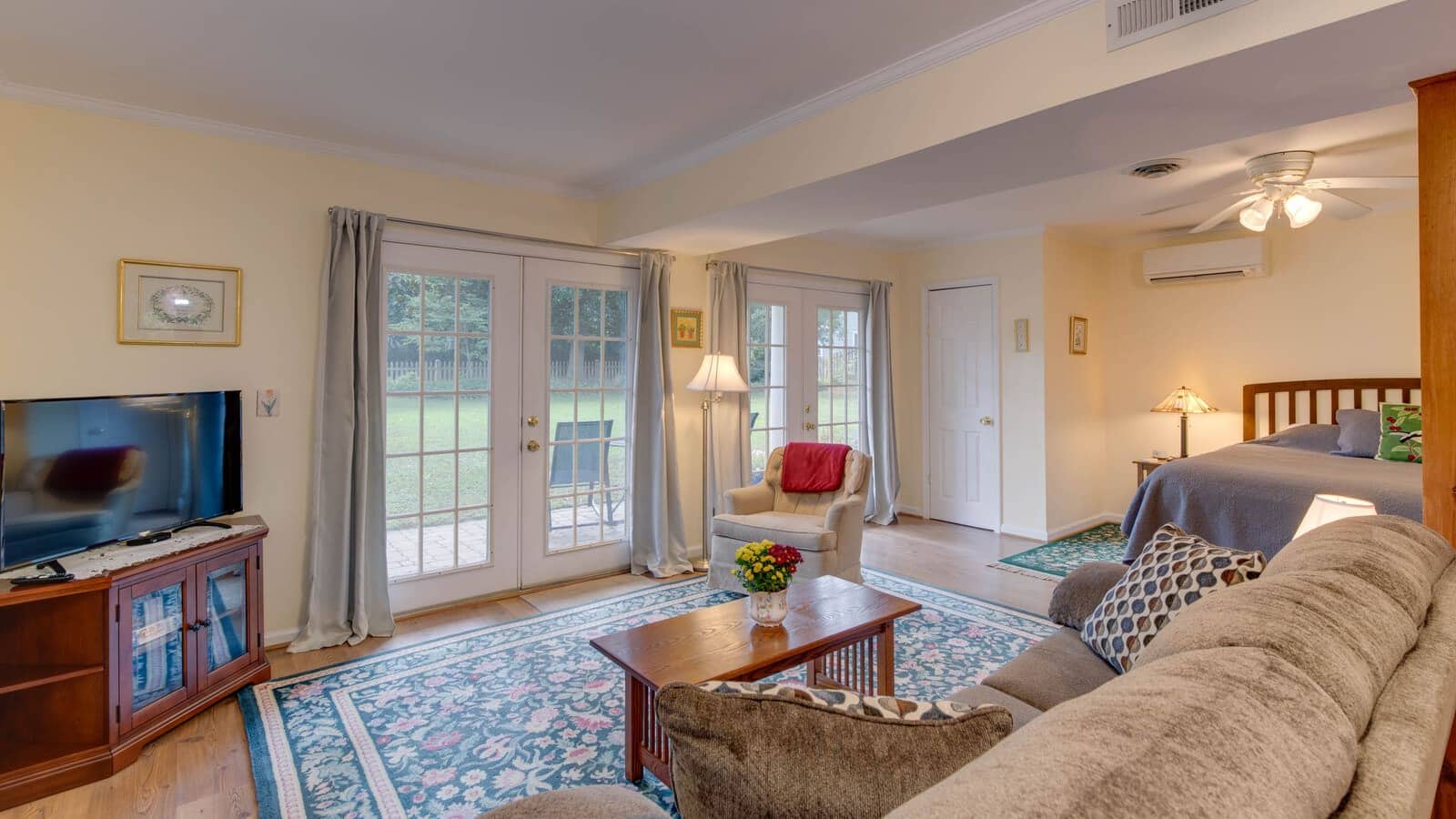 Bedroom suite with cream walls, hardwood flooring, wooden bed with gray bedding, upholstered sofa and chair, wood coffee table, and doors to patio