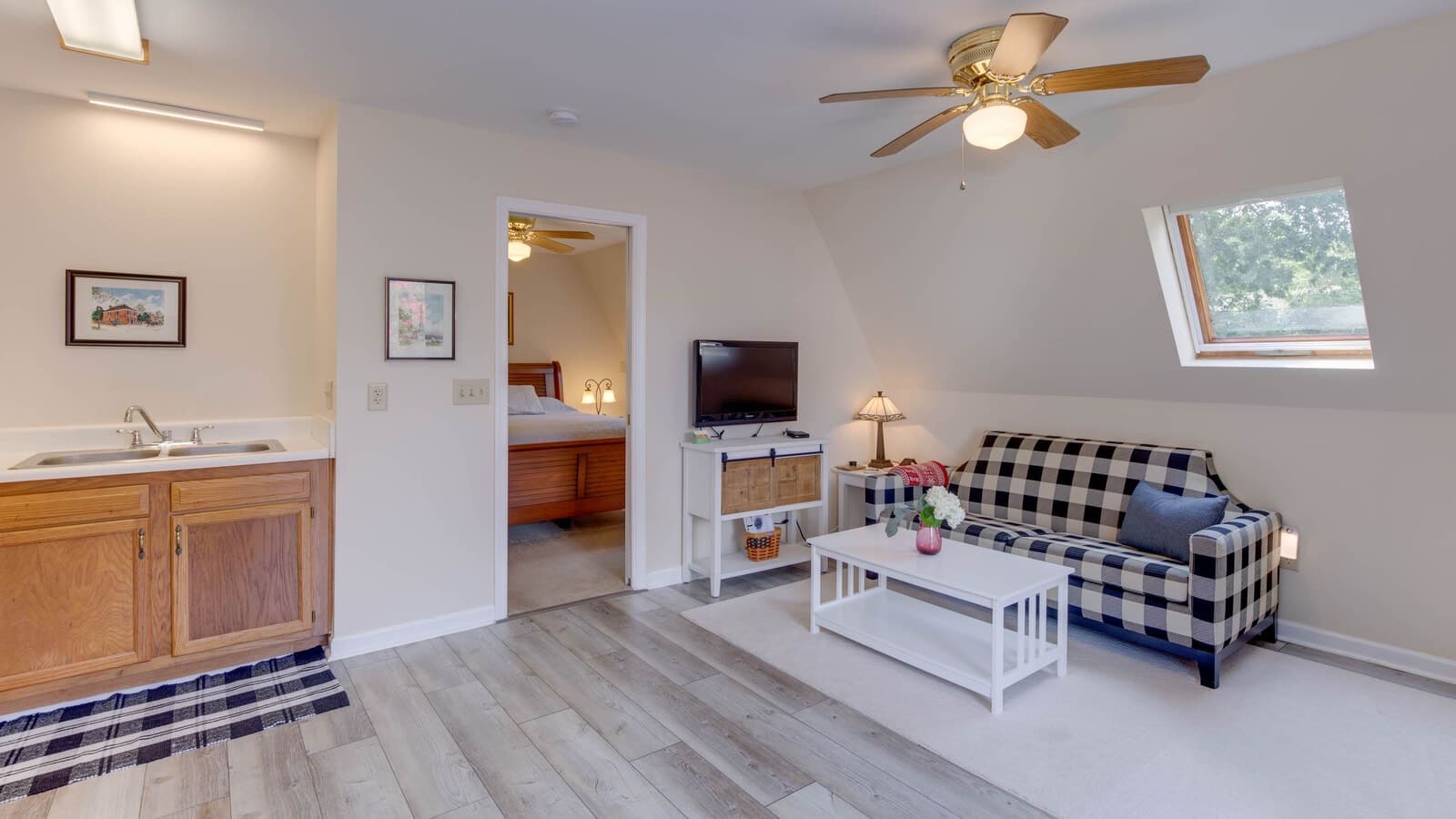 Suite with light colored walls, hardwood flooring, buffalo black and white check upholstered sofa, white coffee table, kitchen sink with wood cabinets, and view into bedroom