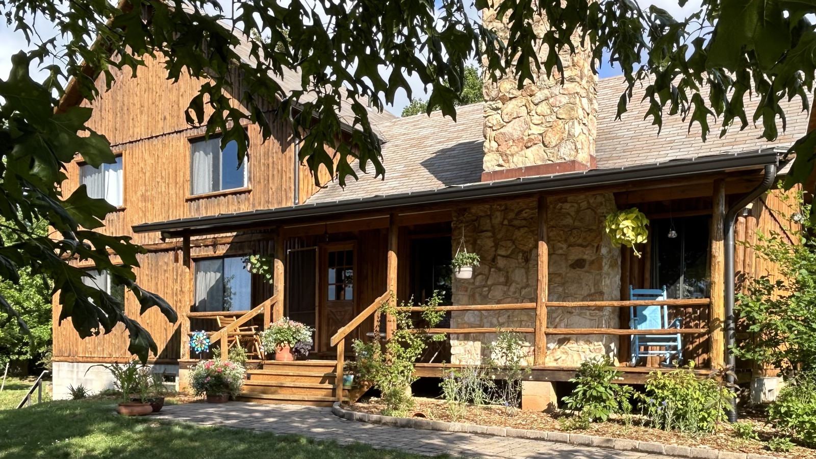 Exterior view of property with wooden siding, stone brick fireplace, and front porch surrounded by green grass, shrubs, and trees