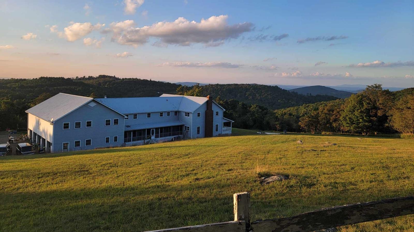 Exterior view of property painted gray with white trim surrounded by large field of grass and rolling hills with green trees in the background