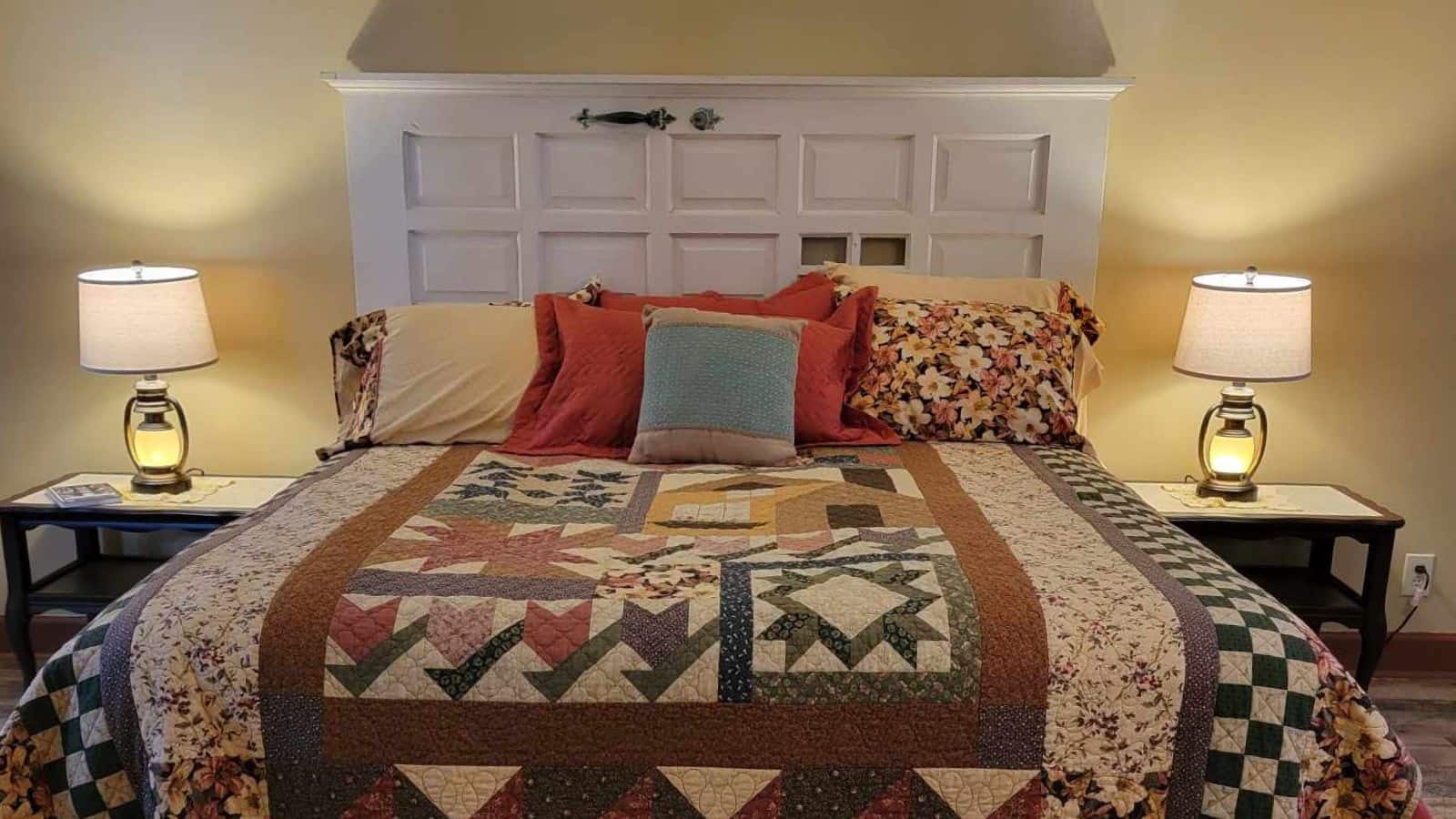 Bedroom with light yellow walls, hardwood flooring, white wooden headboard, multicolored quilt, and matching wooden nightstands with lamps