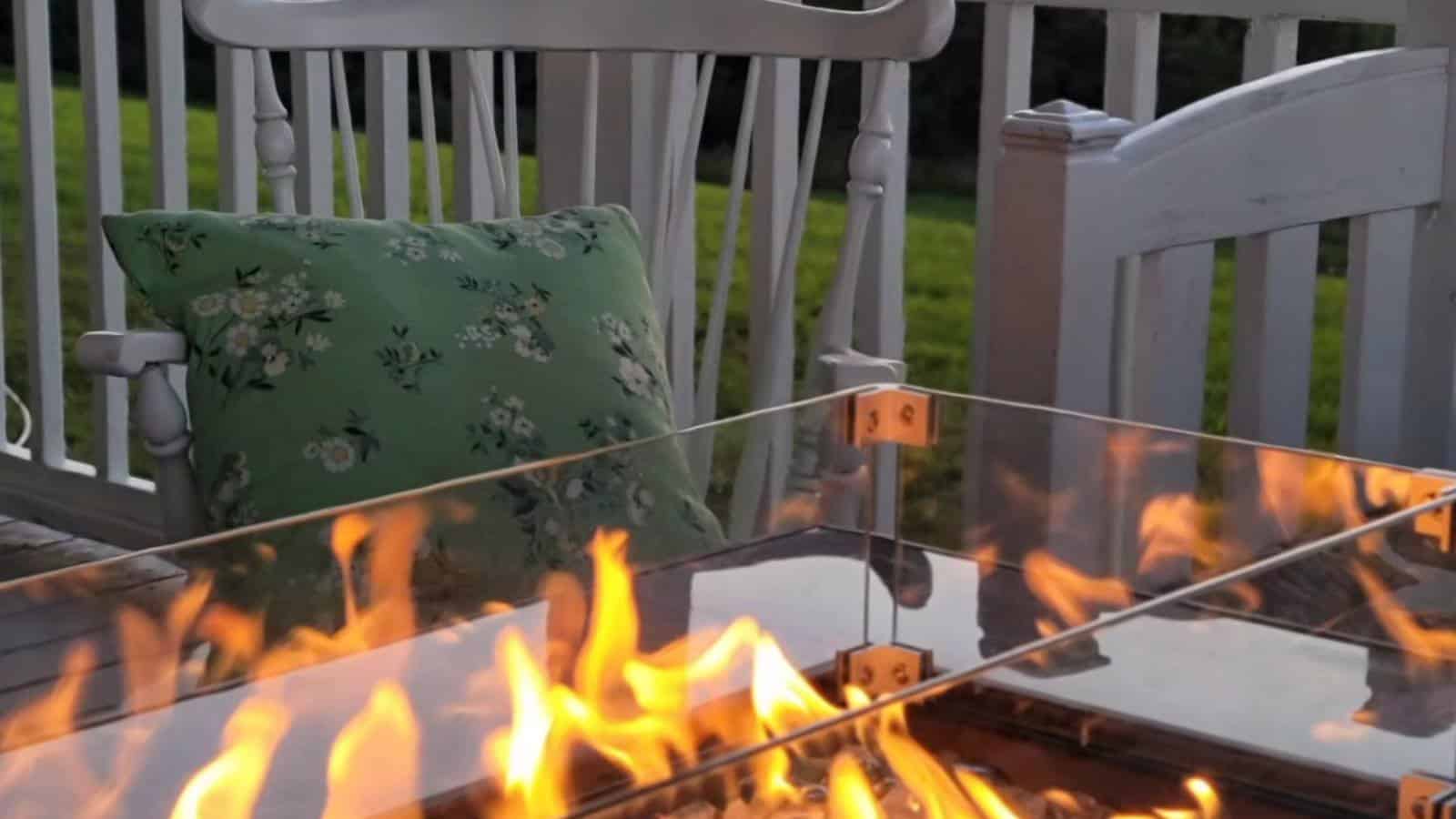 Close up view of gas firepit with glass shield and white rocking chair with green floral pillow