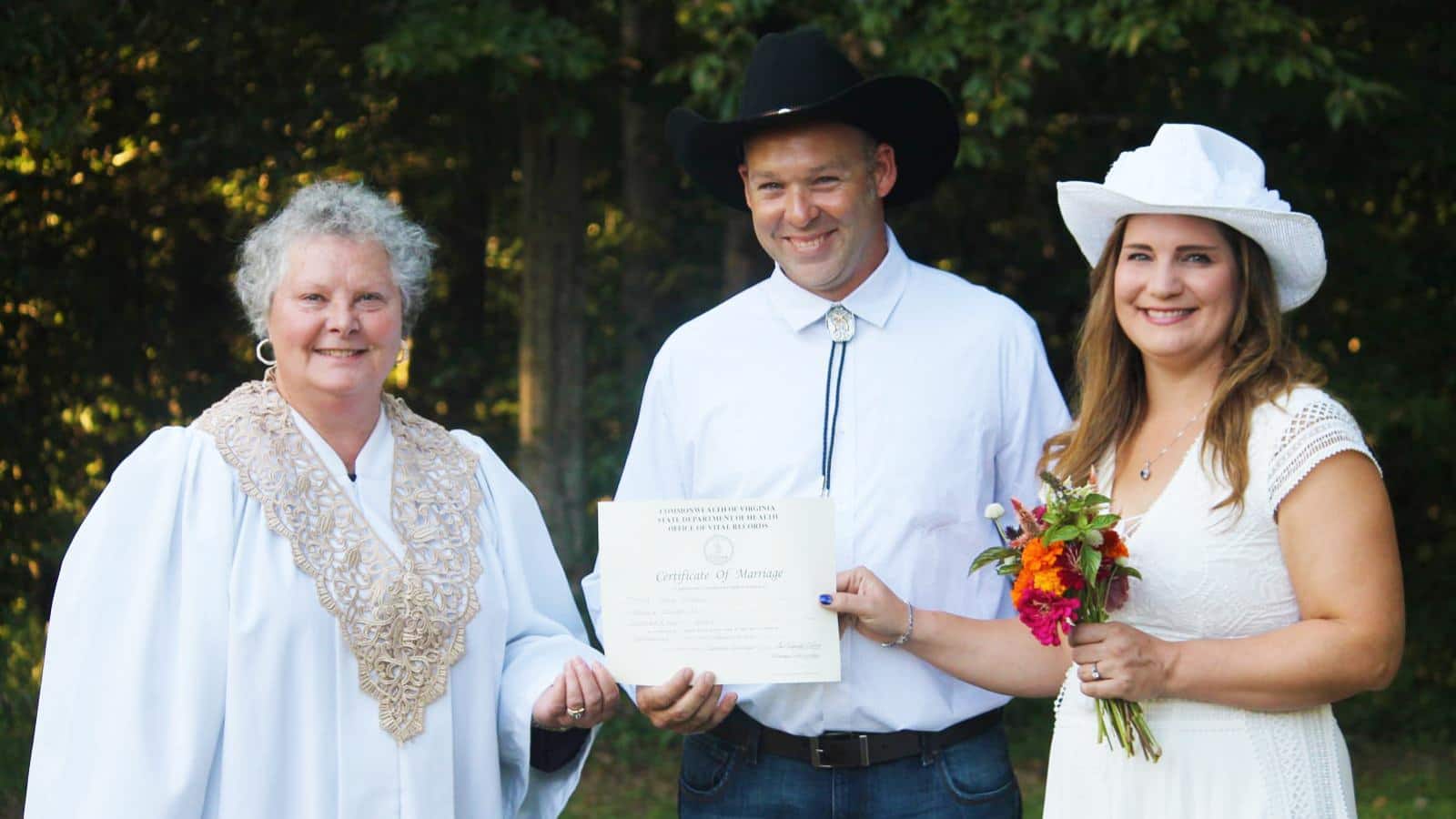 Woman in white robe handing a marriage certificate to a man in a white shirt and black hat and a woman in white hat and white dress