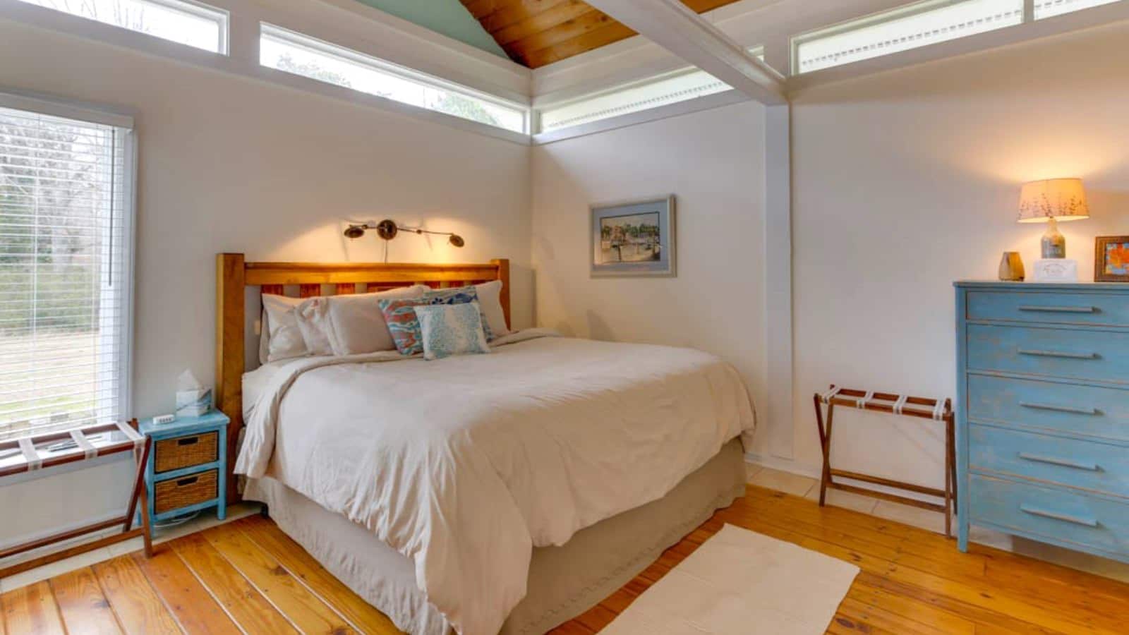 Bedroom with white walls, hardwood flooring, wood bed, light colored bedding, and teal wooden dresser