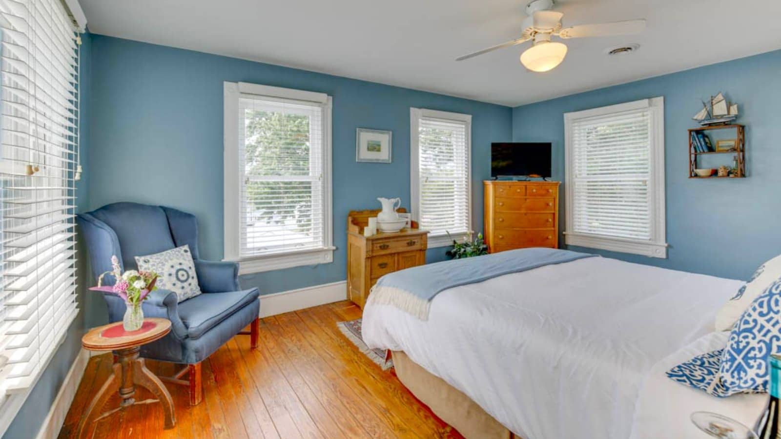 Bedroom with blue walls, hardwood flooring, bed with white bedding, blue upholstered armchair, and wooden dresser
