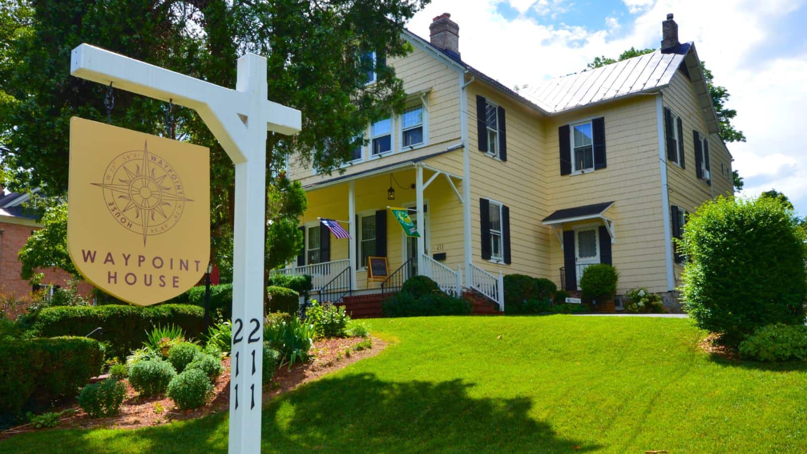 Exterior view of property painted yellow with white trim and black shutters surrounded by green grass, shrubs, gardens, and trees